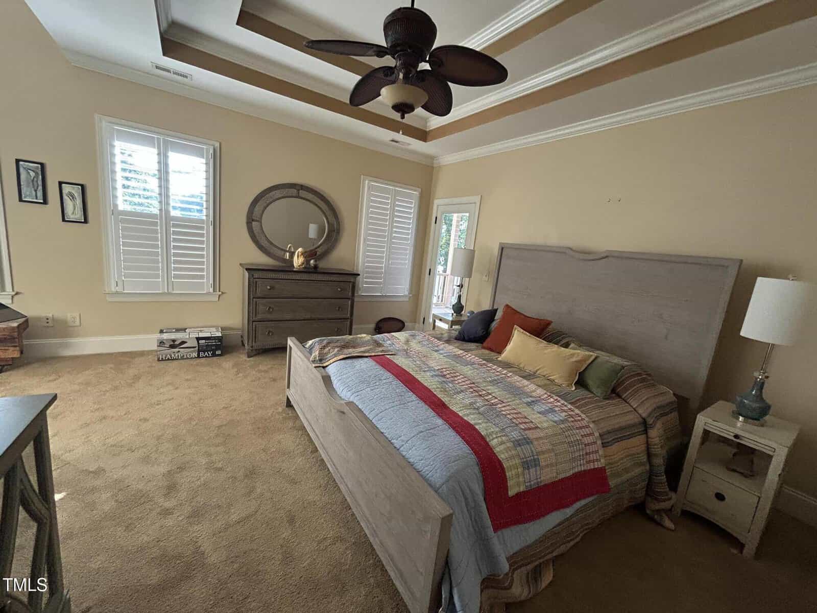 A bedroom with a wooden bed, colorful quilt, dresser with a round mirror, table lamp, ceiling fan, carpeted floor, shuttered windows, and framed wall art. A door leads outside. Boxes are stacked near the dresser.