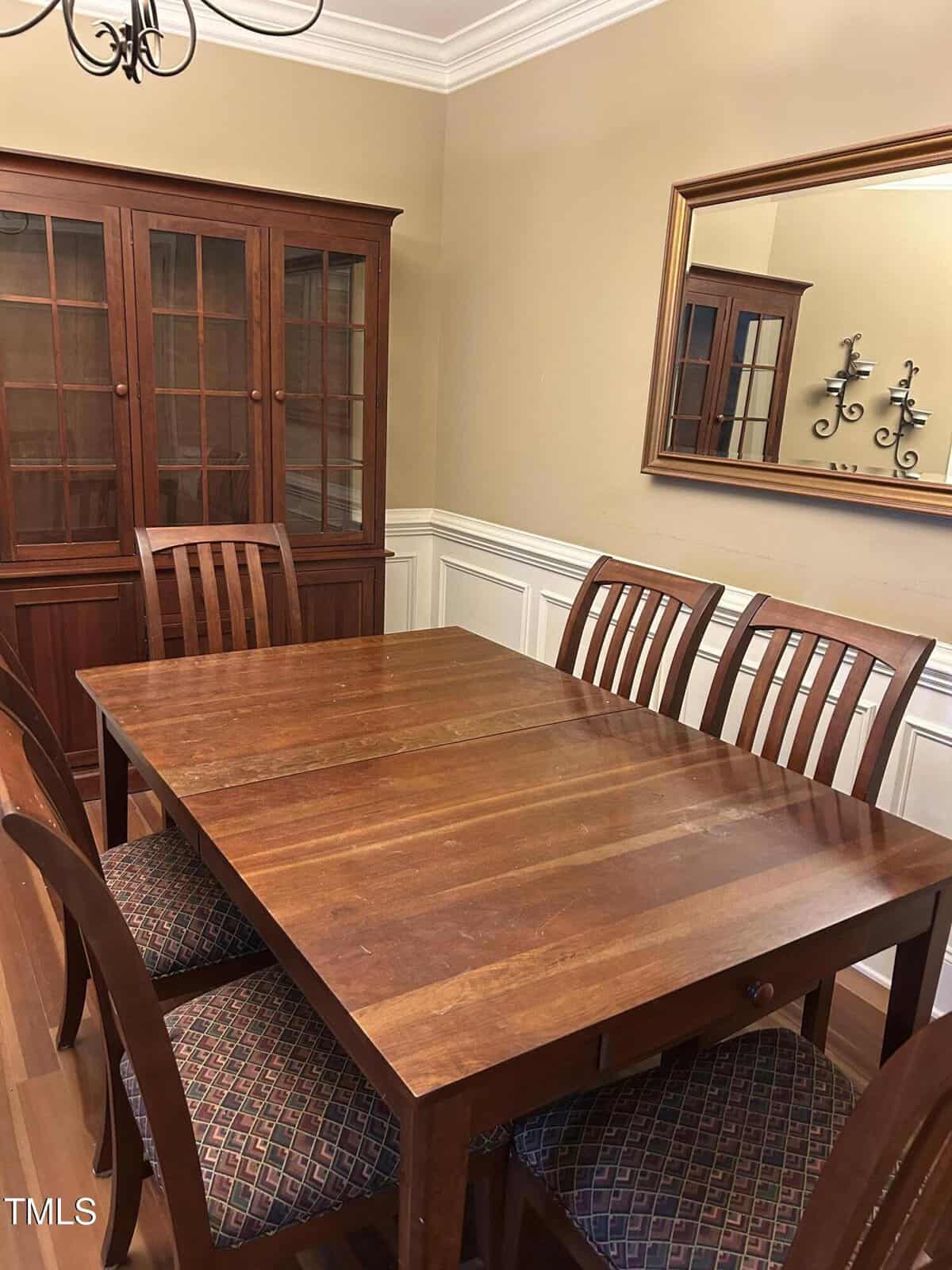 A wooden dining table with six matching chairs sits in a room with beige walls, white wainscoting, a china cabinet, a large mirror, and a wall sconce. The chairs have patterned fabric seats.