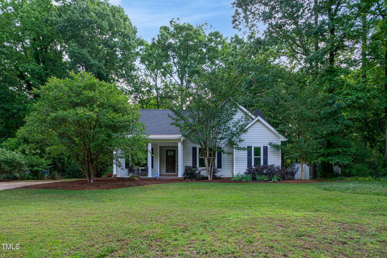 A small white house with a front porch sits surrounded by large leafy trees and lush green grass, creating a peaceful, secluded suburban setting under a clear blue sky.
