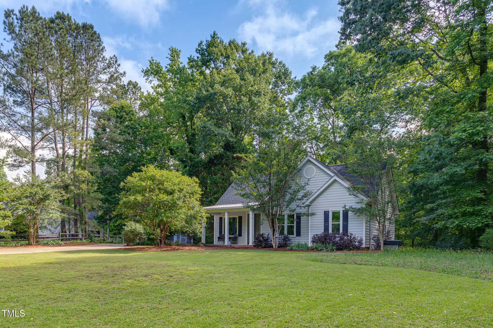 A small white house with a front porch sits surrounded by tall trees and lush greenery, with a large grassy yard in the foreground under a blue sky with scattered clouds.