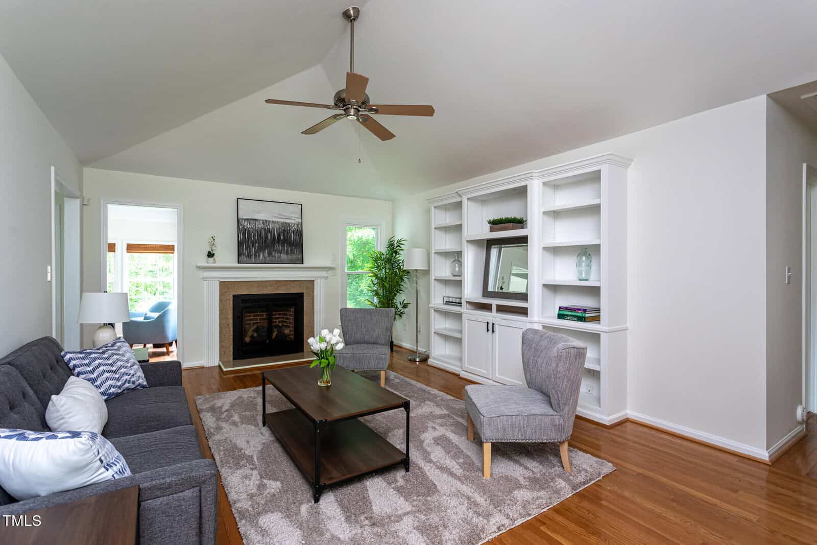 Bright living room with a vaulted ceiling, ceiling fan, gray sofas, armchair, wooden coffee tables, built-in white shelves, fireplace with art above, hardwood floors, and natural light from nearby windows and doors.