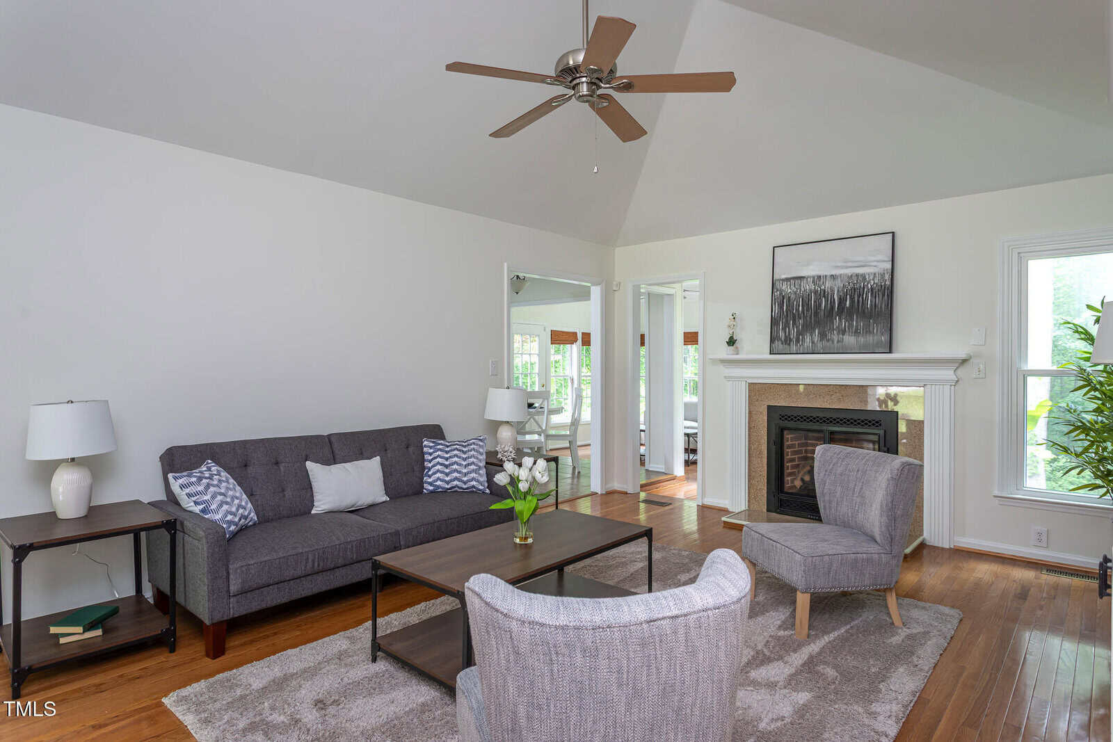 A bright living room with gray furniture, a coffee table, fireplace, and ceiling fan. Large windows let in natural light, and artwork and a plant add decorative touches.