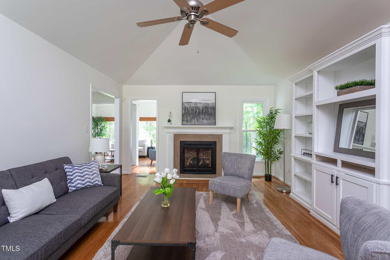 Modern living room with gray sofas, a wooden coffee table with white flowers, a fireplace, wall art, and a ceiling fan. Built-in white shelves and hardwood floors complete the bright, airy space.