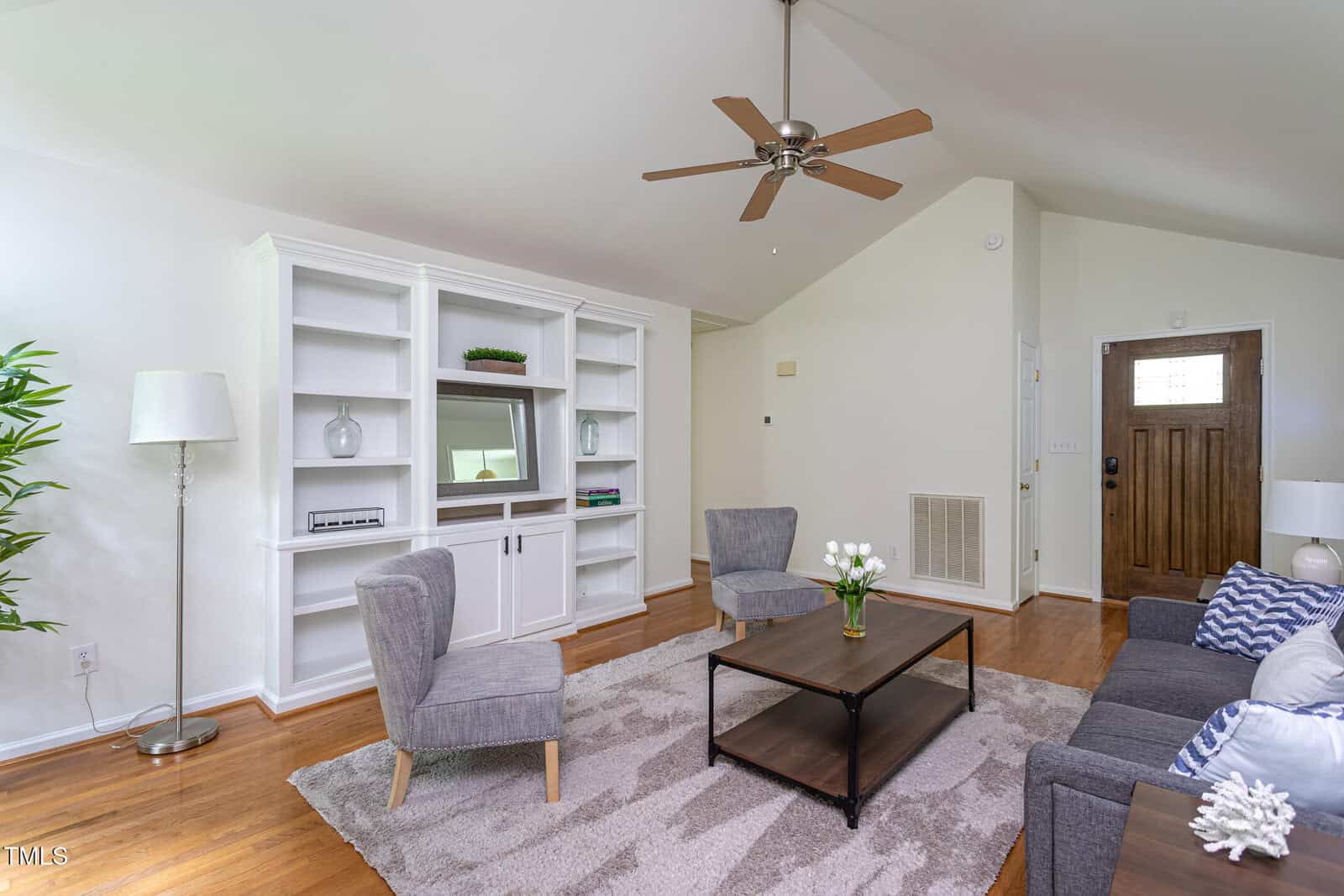 Living room with two gray armchairs, a gray sofa, a wooden coffee table, a floor lamp, a built-in white shelving unit with a mirror, a ceiling fan, houseplant, and a wooden front door. Hardwood floors and a beige rug.