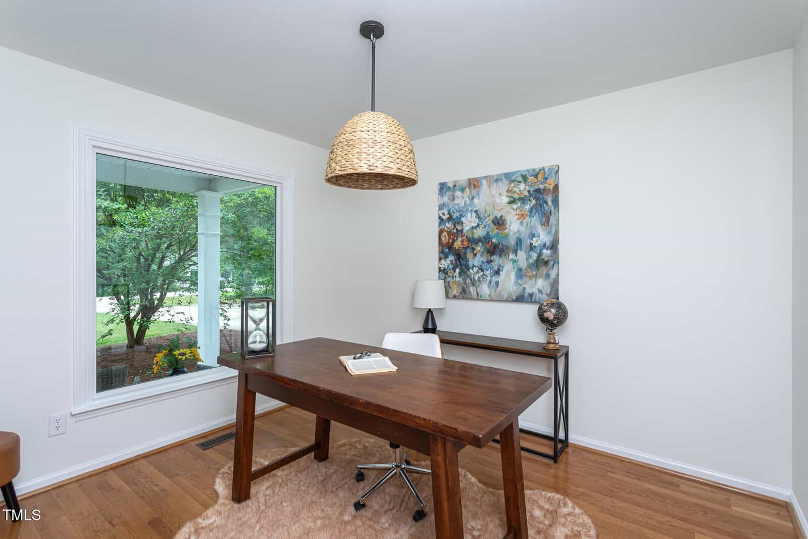A home office with a wooden desk, rolling chair, large window, woven pendant light, abstract wall art, side table with a lamp and globe, and a furry rug on hardwood floors. Trees are visible outside the window.