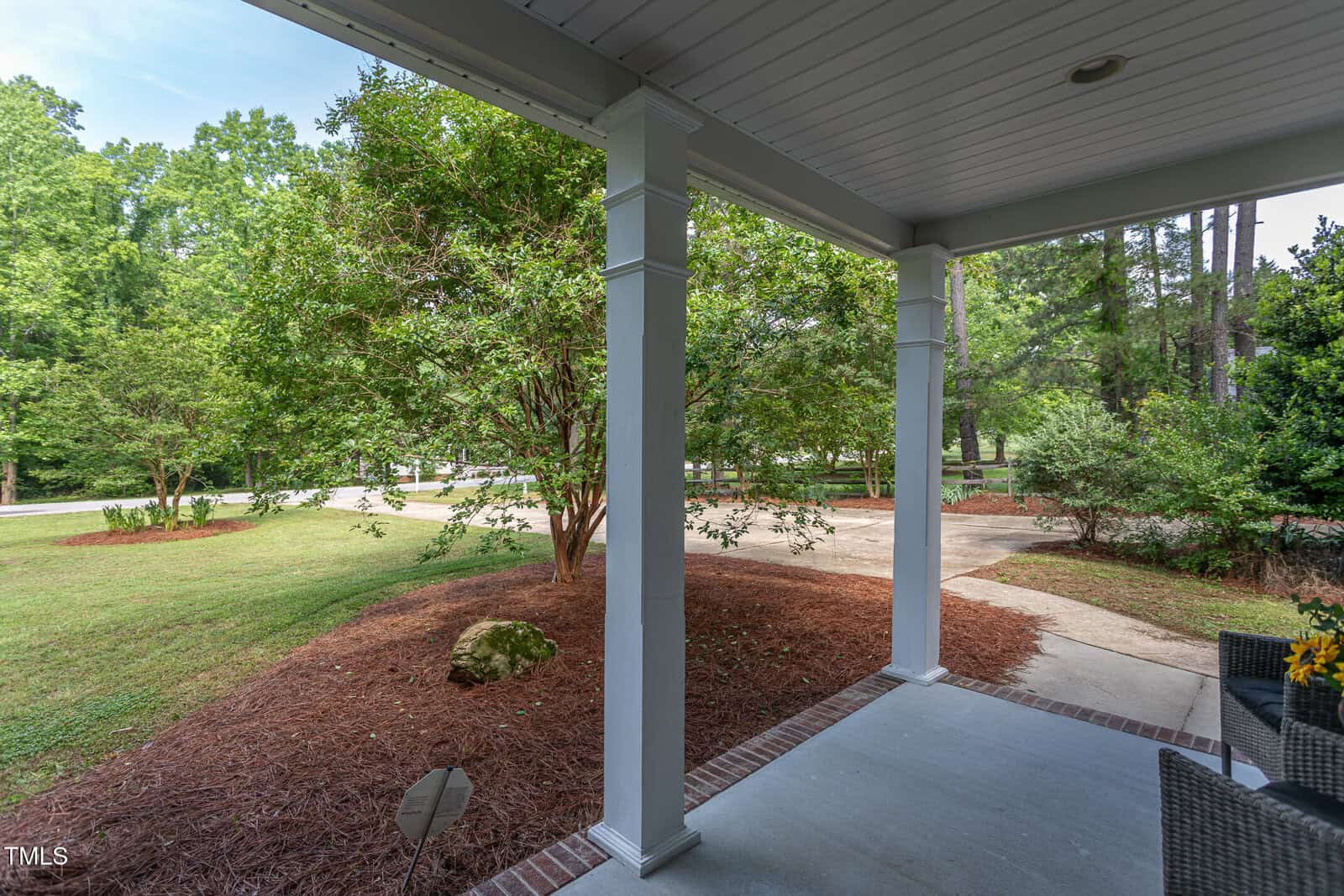 Covered front porch with white columns overlooking a landscaped yard with trees, mulch, and a curved driveway. A wicker chair with a yellow pillow and potted plant are visible on the porch.