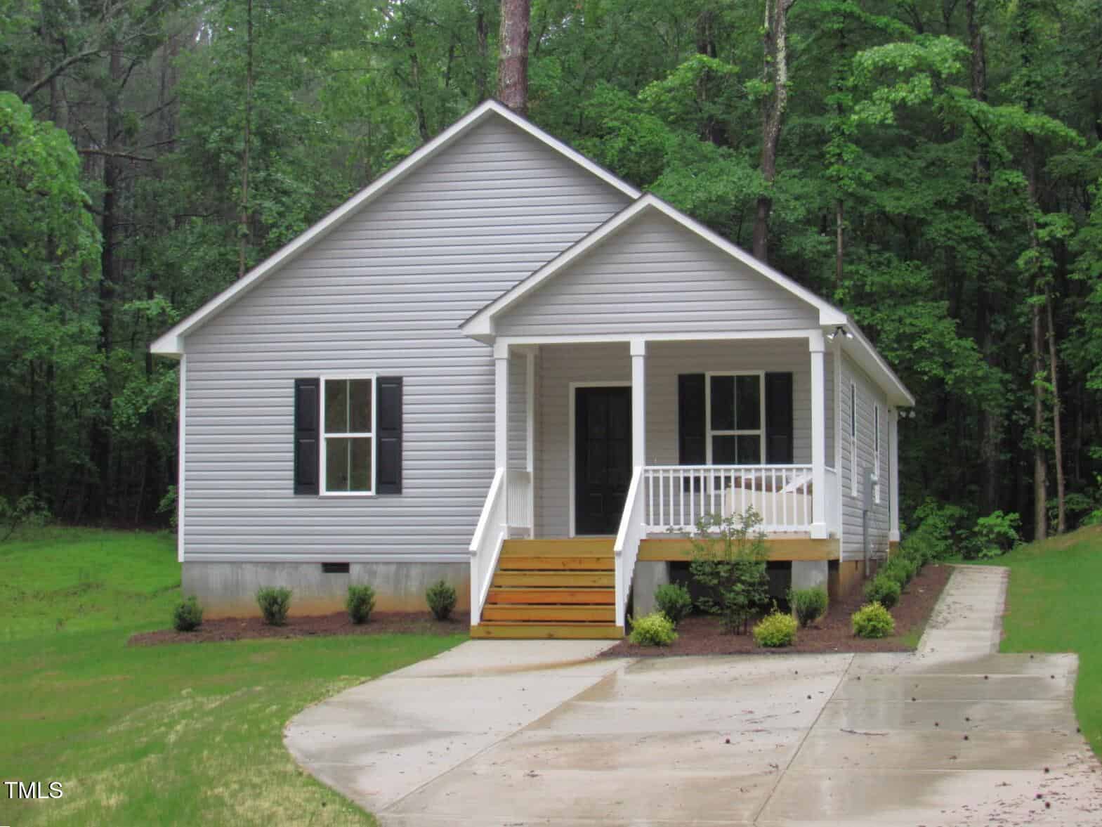 A small, single-story gray house with white trim, a front porch with steps and railing, black shutters, and a curved concrete driveway, surrounded by green trees and lawn.