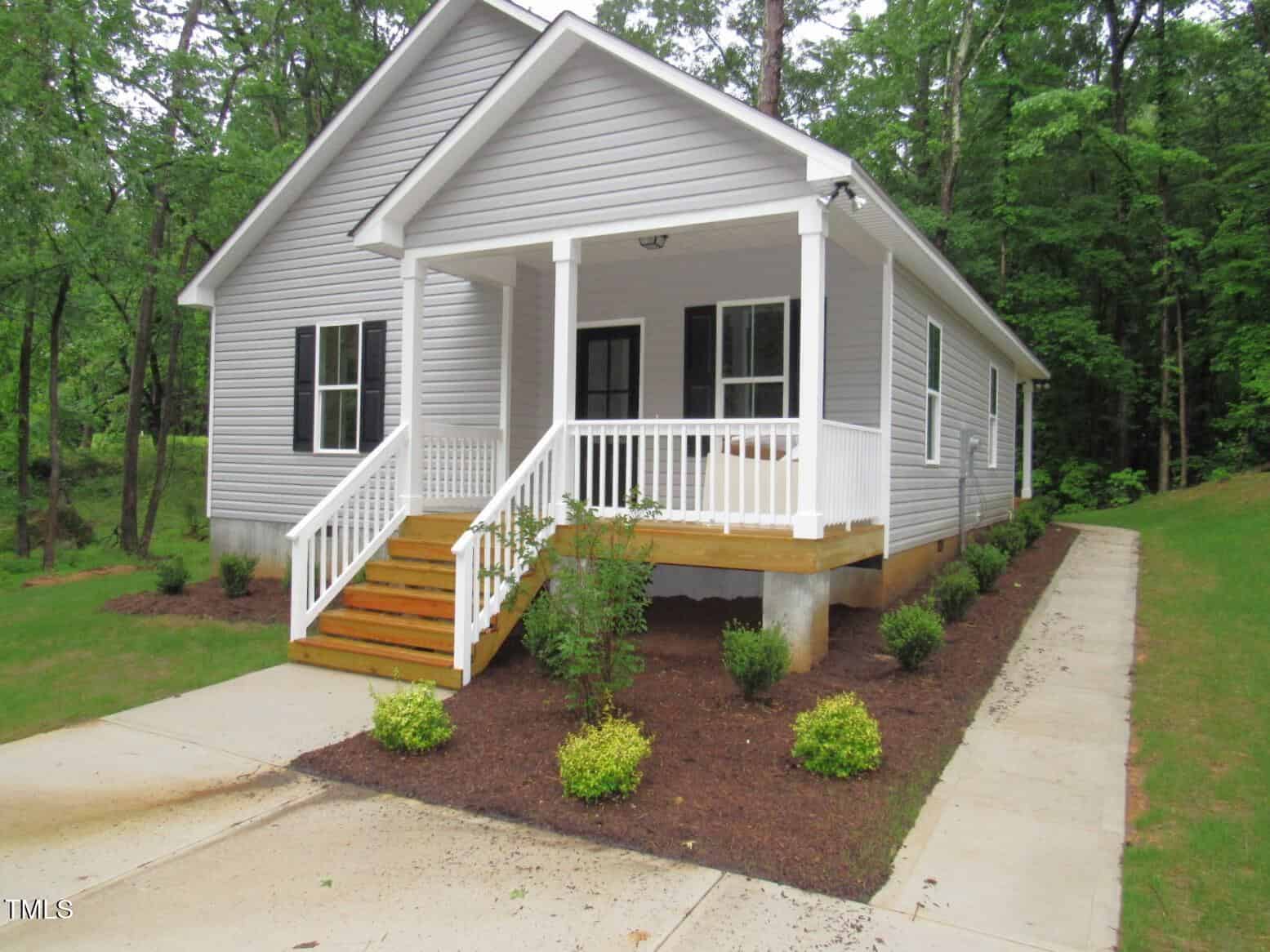A small gray house with white trim, a covered front porch, and wooden steps, surrounded by green shrubs and trees. A concrete sidewalk leads to the front and along the side of the house.