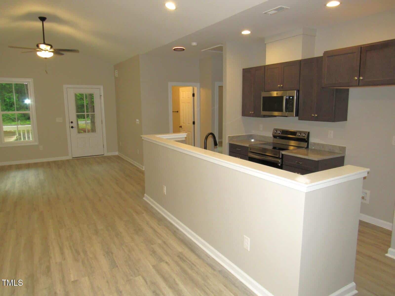 Open-concept kitchen and living area with light wood floors, neutral walls, stainless steel appliances, dark cabinets, and a ceiling fan. A door and large window provide natural light at the far end of the room.