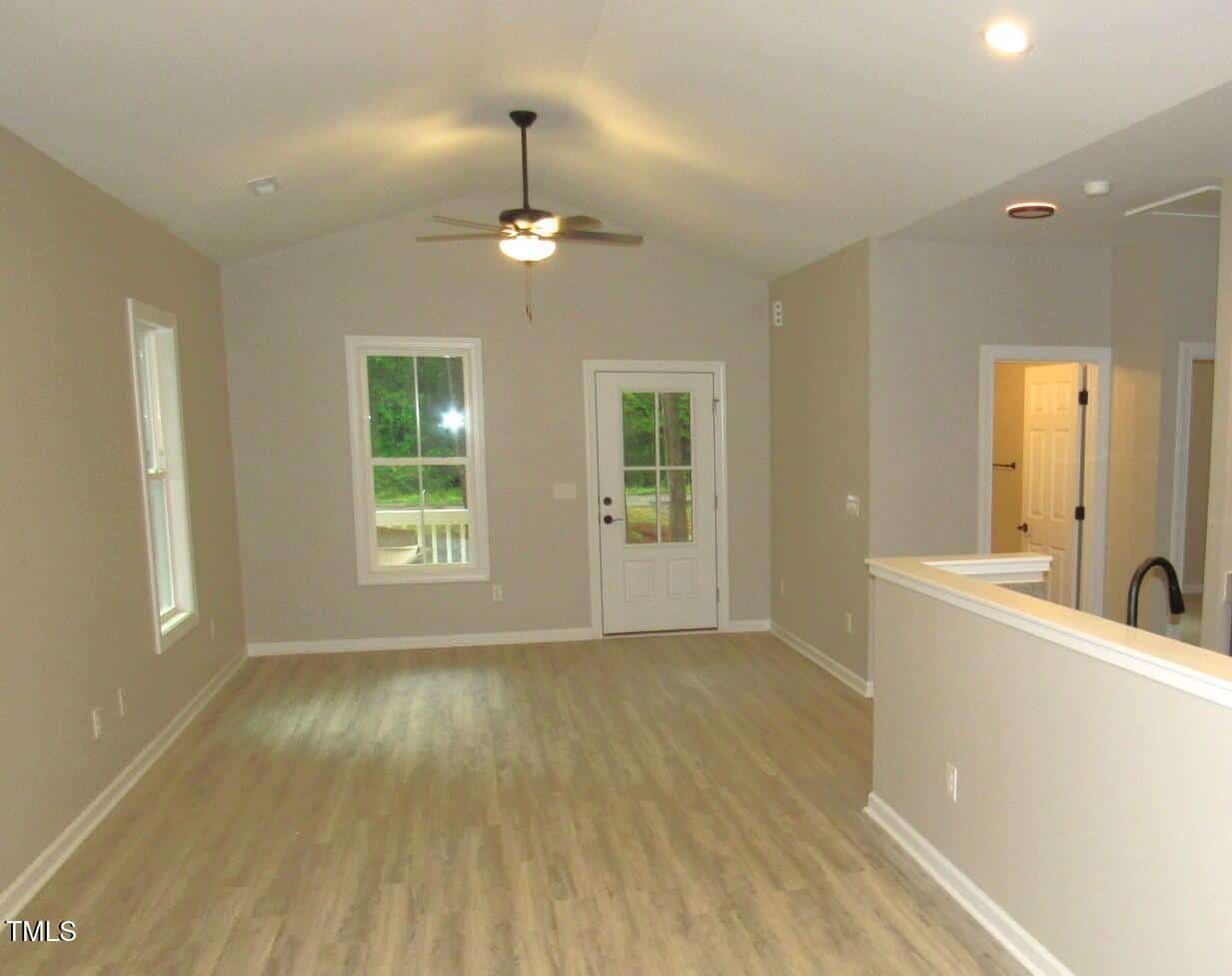 A bright, empty room with light wood flooring, light gray walls, a ceiling fan, two windows, a glass door, and an open doorway leading to another area of the house.