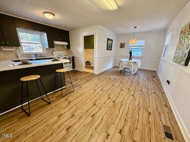 A kitchen with dark cabinets, white appliances, and a breakfast bar with two stools is shown. The kitchen opens into a dining area with a small round table and chairs by a window. The floor is wood-patterned.