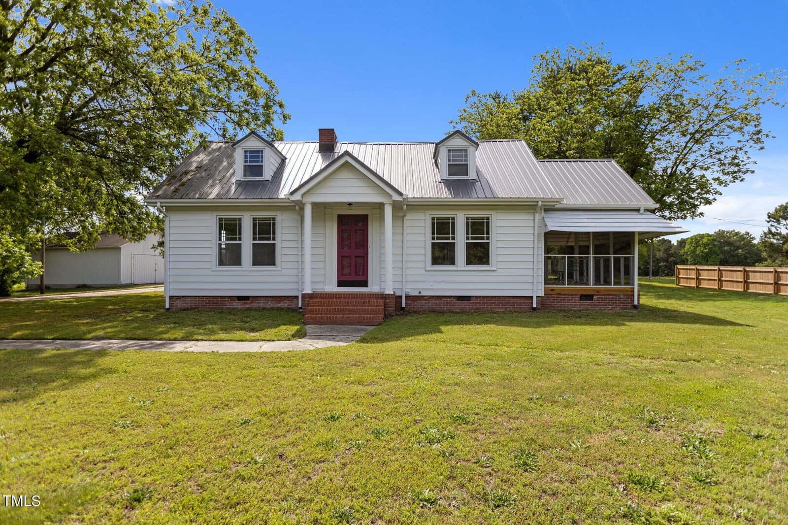 A charming single-story white house at 2227 US-15, Creedmoor, featuring a red front door, metal roof, two dormer windows, brick steps, a screened porch to the right, and a large green lawn under a clear blue sky.