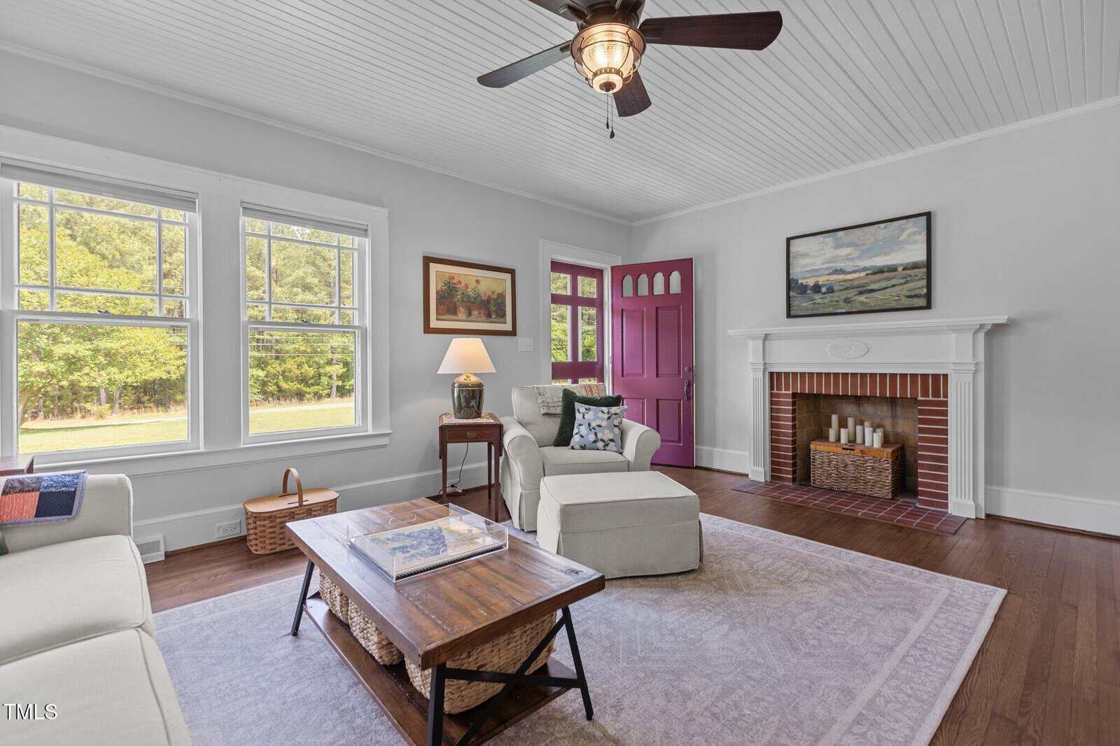 A bright living room with white walls, wood floors, and a white ceiling. It has a brick fireplace, a purple front door, a ceiling fan, comfortable seating, and a wooden coffee table on a light rug.