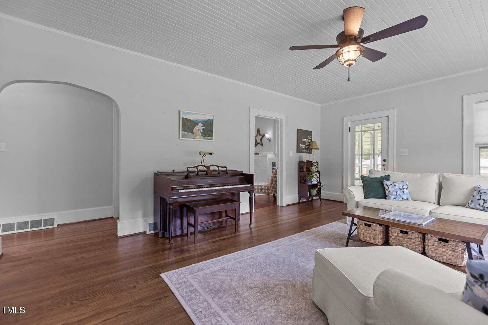 A living room with hardwood floors, a ceiling fan, an upright piano against one wall, a coffee table, a sectional sofa with cushions, and a light purple rug. There are two doorways and several pieces of art on the walls.