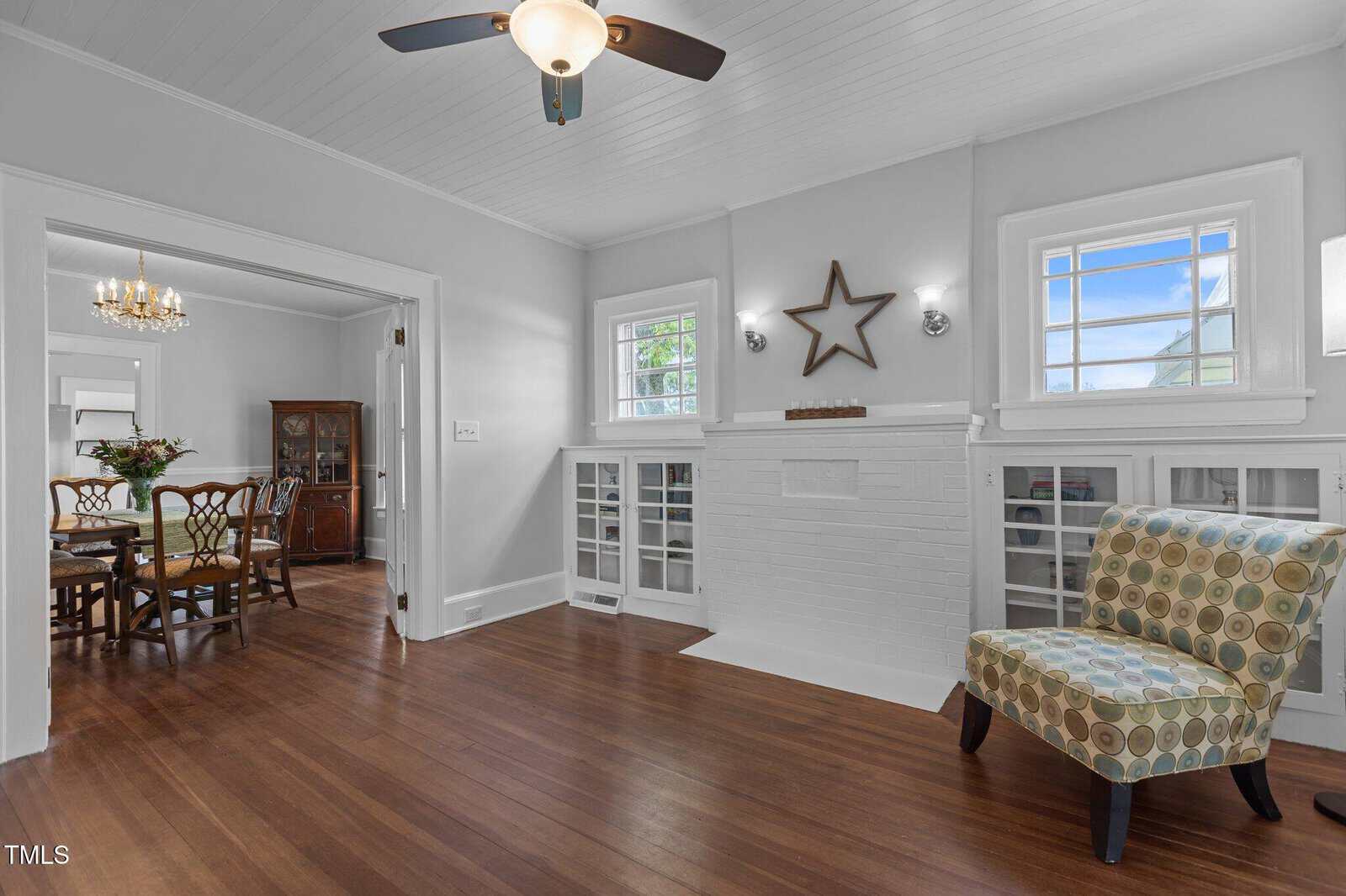 A bright living room with hardwood floors, white walls, a ceiling fan, built-in cabinets with glass doors, a patterned armchair, and a white brick fireplace. A dining area is visible through an open doorway.
