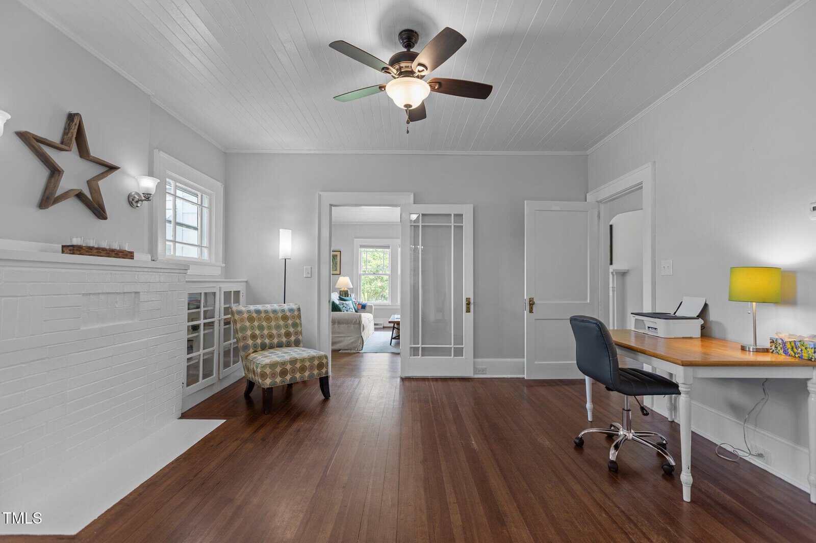 A bright room with white walls and ceiling, hardwood floors, a ceiling fan, a desk with a yellow lamp, a patterned armchair, a decorative star on the wall, and glass doors leading to another room.