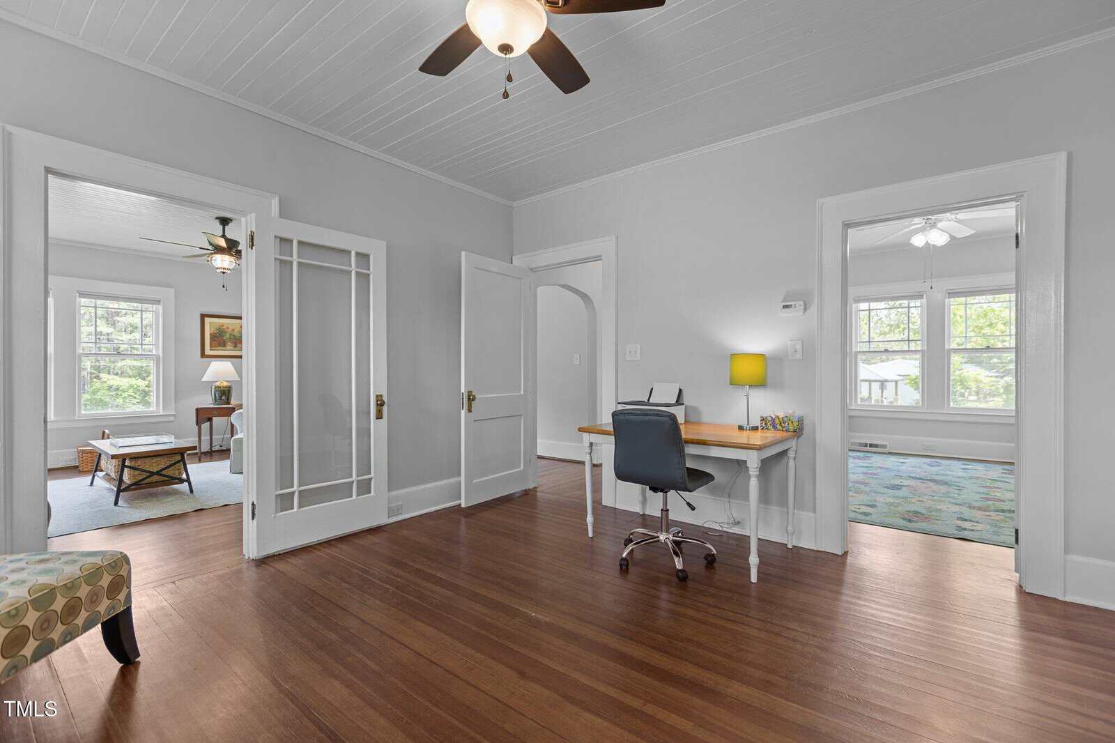 A home office with a wooden floor, a desk and chair, ceiling fan, and white walls. Two adjoining rooms are visible, each with large windows and ceiling fans, and a patterned chair sits in the foreground.