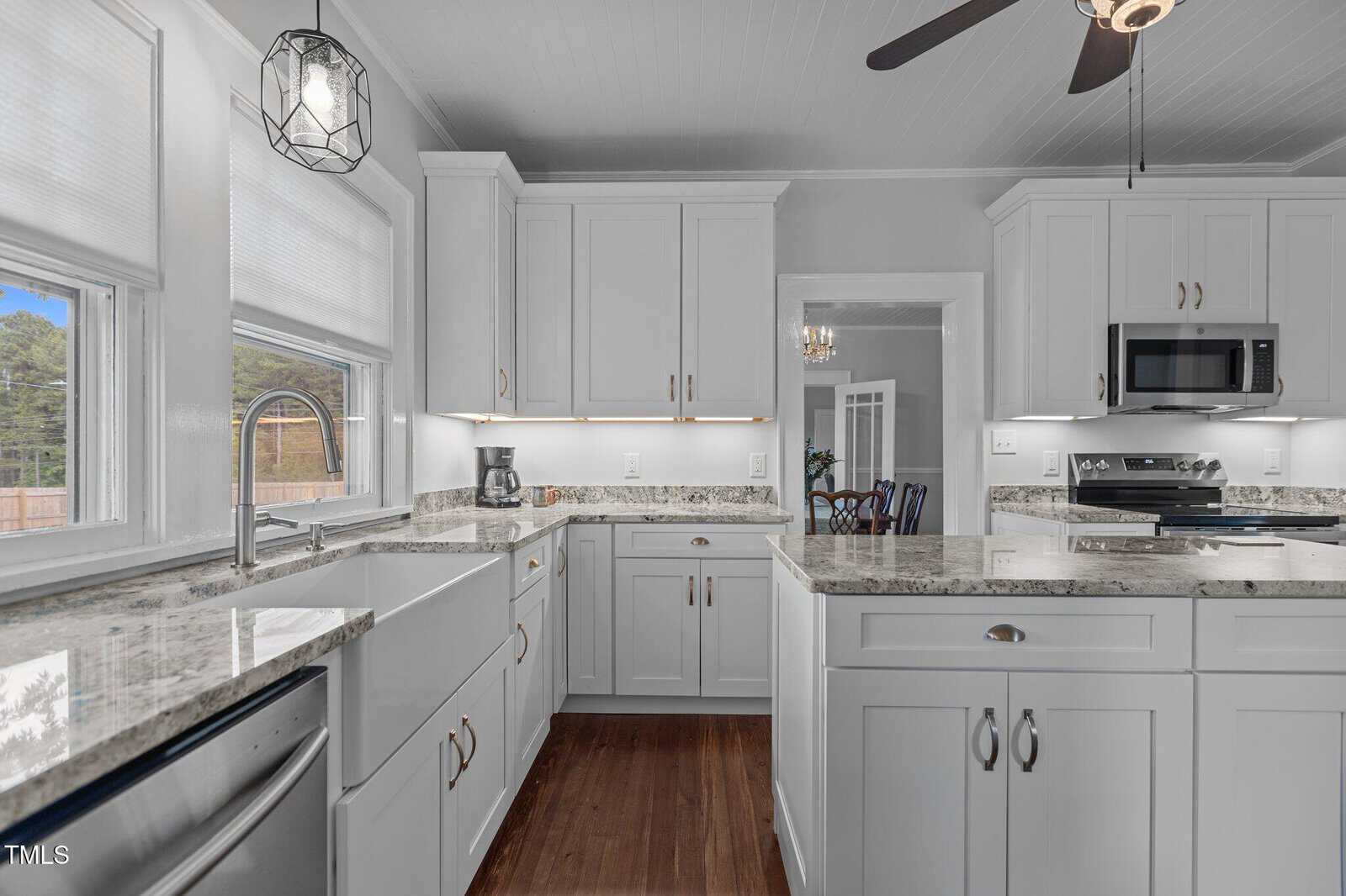 A modern kitchen with white cabinets, granite countertops, a farmhouse sink, stainless steel appliances, and wood floors. A window above the sink lets in natural light, and a ceiling fan hangs above.