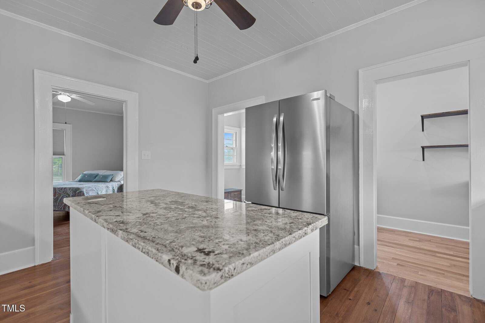 A modern kitchen with a marble island countertop, stainless steel refrigerator, and hardwood floors. Doorways lead to a bedroom, a room with shelves, and another area with a window.