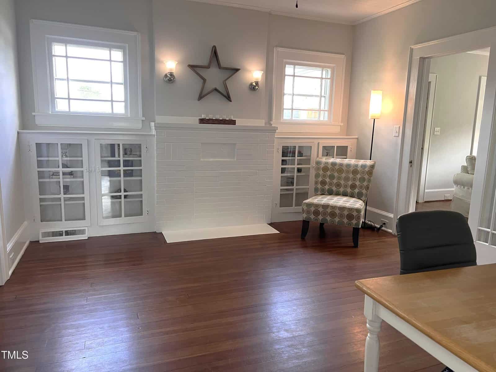 A bright room with hardwood floors, white walls, a white fireplace with glass-fronted built-in shelves, a patterned armchair, a desk, and two windows letting in natural light. A star decoration hangs above the fireplace.