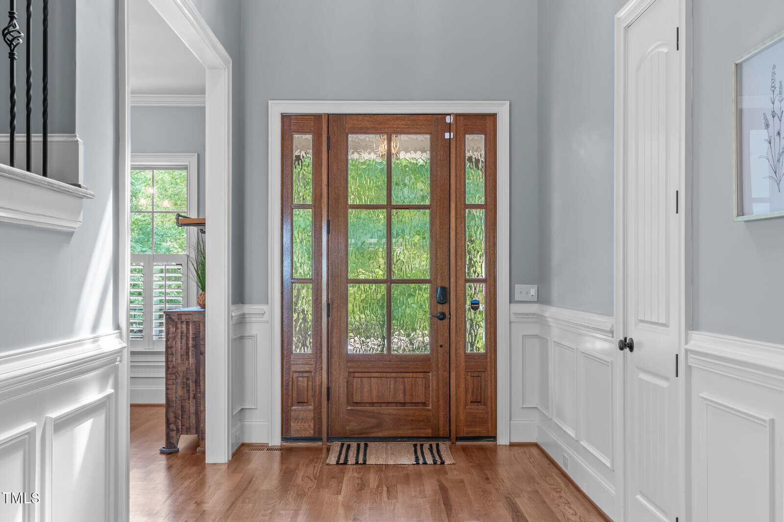 A bright entryway with light gray walls, white wainscoting, and a wooden front door with glass panels. Natural light enters through the door, and a small striped rug is placed on the wood floor.