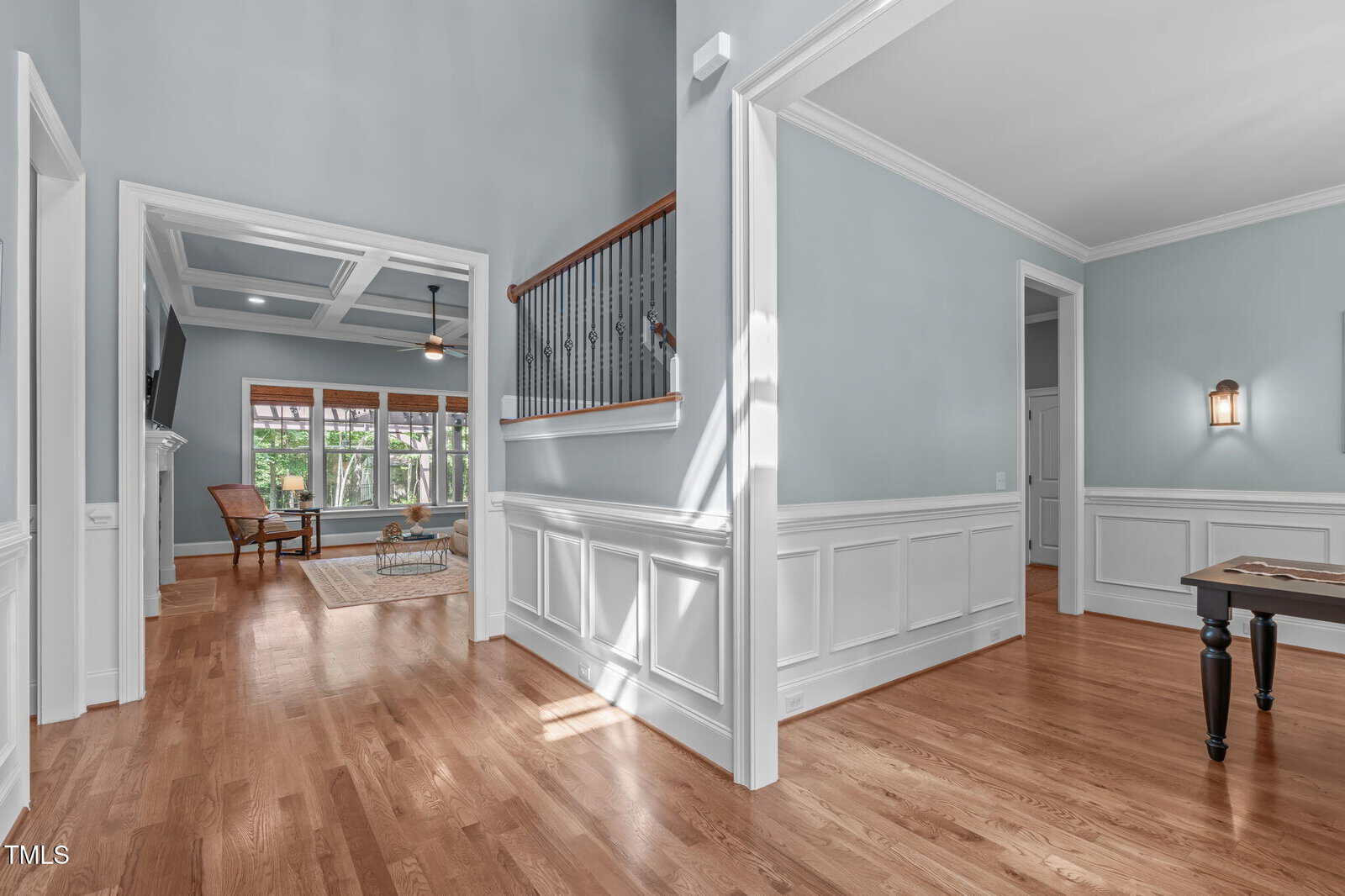 A hallway with light blue walls, white wainscoting, and wood floors leads to an open living area with large windows, a ceiling fan, and a wooden chair. Natural light enters through the windows.