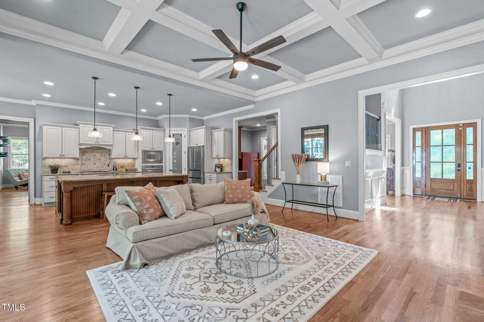 Spacious open-concept living area with a beige sofa and patterned rug, coffered ceiling, ceiling fan, and view of the kitchen with white cabinets, island, pendant lights, and entryway with wooden double doors.