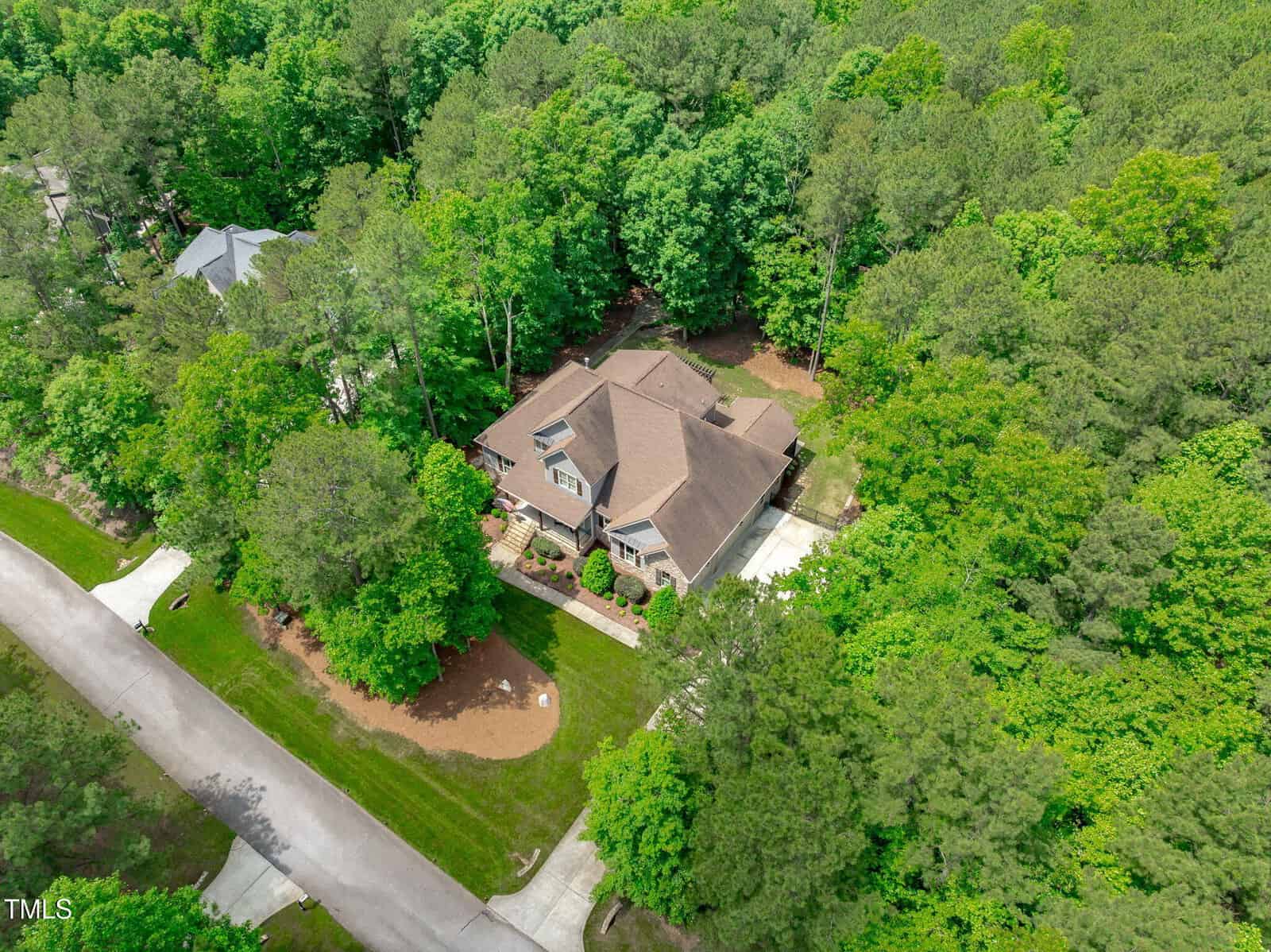Aerial view of a two-story house with a brown roof, surrounded by trees and greenery, located in a suburban neighborhood with nearby houses and a paved road.