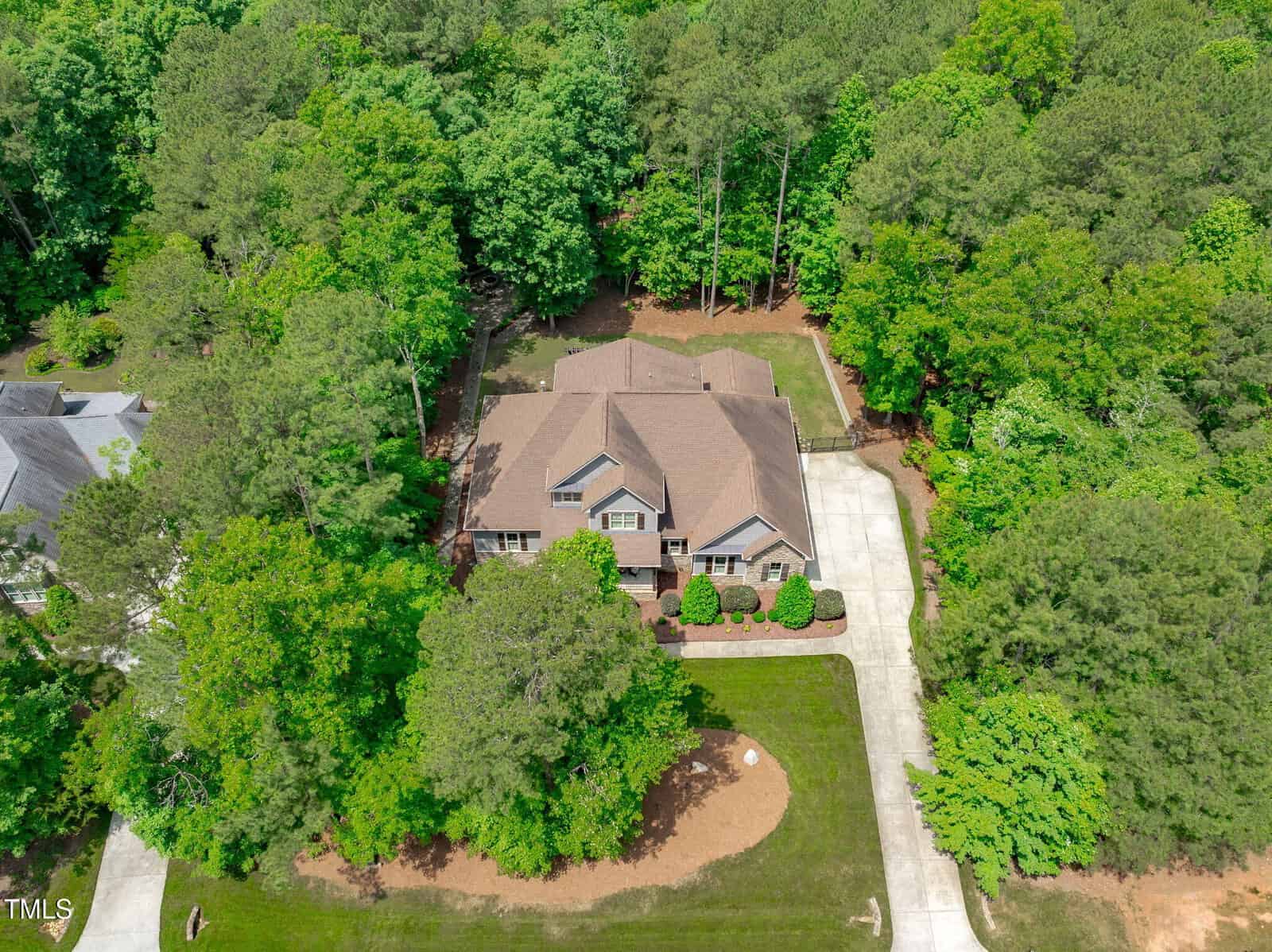 Aerial view of a large house with a brown roof surrounded by tall green trees, a spacious driveway, and a neatly manicured lawn in a wooded residential area.