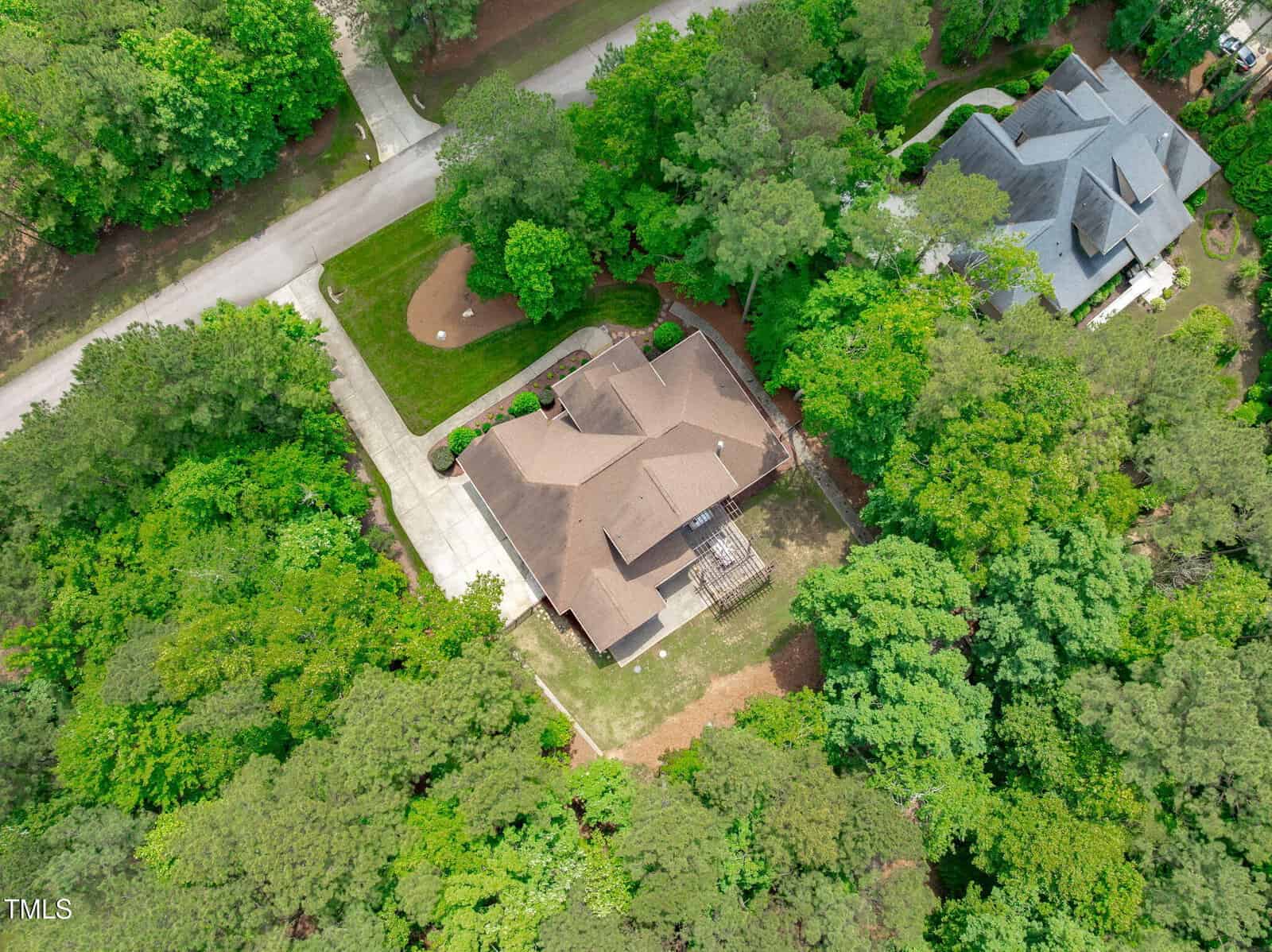 Aerial view of a suburban house with a brown roof, driveway, and surrounding green trees. A road runs in front of the property, and a neighboring house is visible to the right.