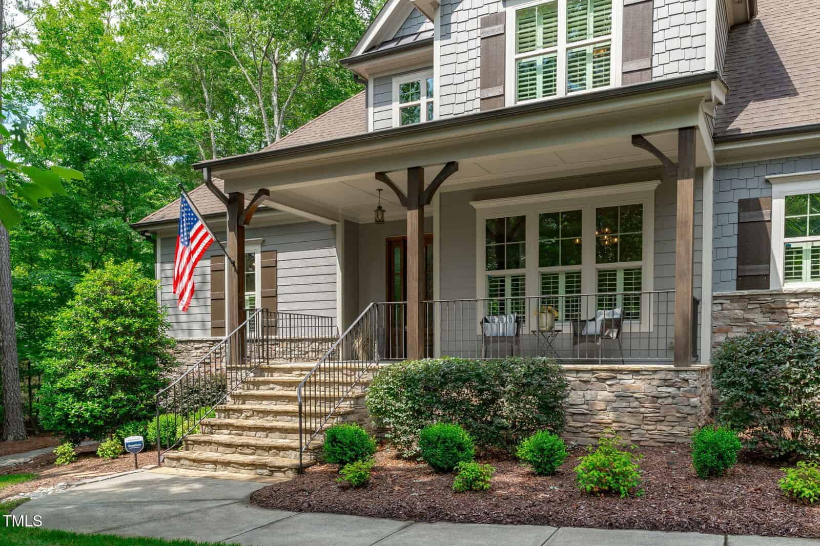 A two-story house with gray siding and stone accents, a covered front porch with white chairs, a metal railing on the steps, and an American flag. The yard has shrubs, mulch, and a concrete walkway.