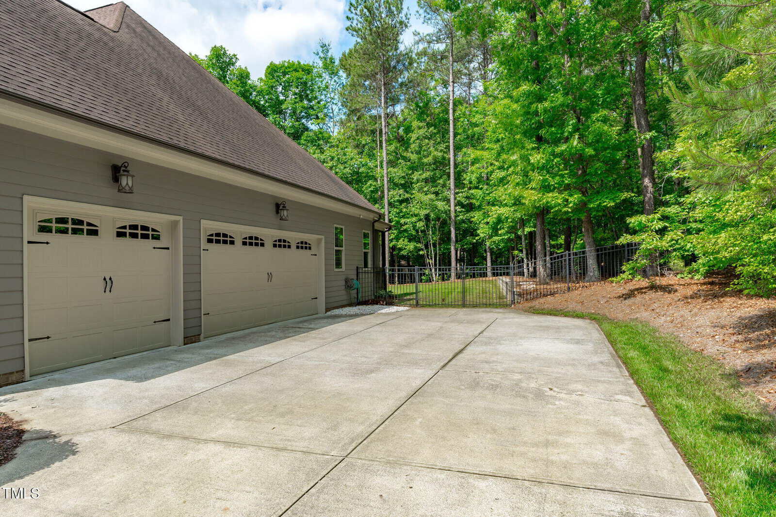 A concrete driveway leads to a two-car garage attached to a gray house. The garage doors have decorative windows, and the area is bordered by green grass, trees, and a black metal fence.
