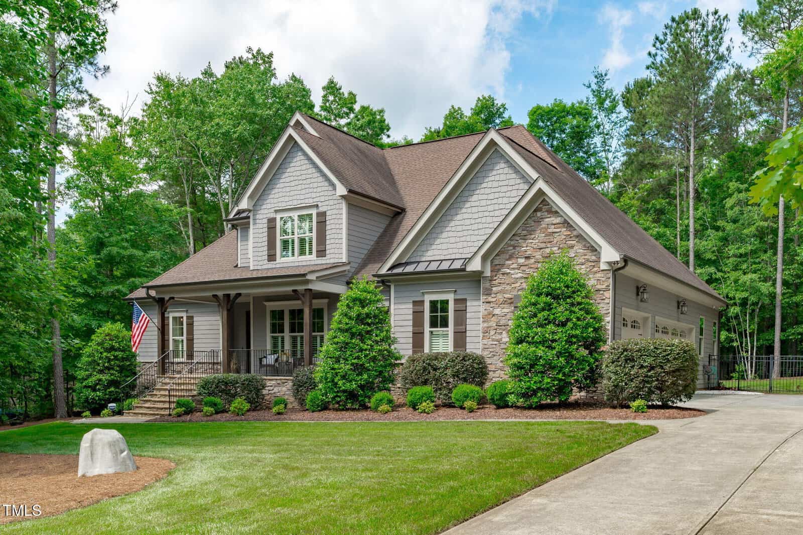A two-story suburban house at 677 Juliette Court in Creedmoor features a stone and siding exterior, a covered front porch, manicured lawn, shrubs, and an American flag. The curved driveway leads to a three-car garage surrounded by trees.