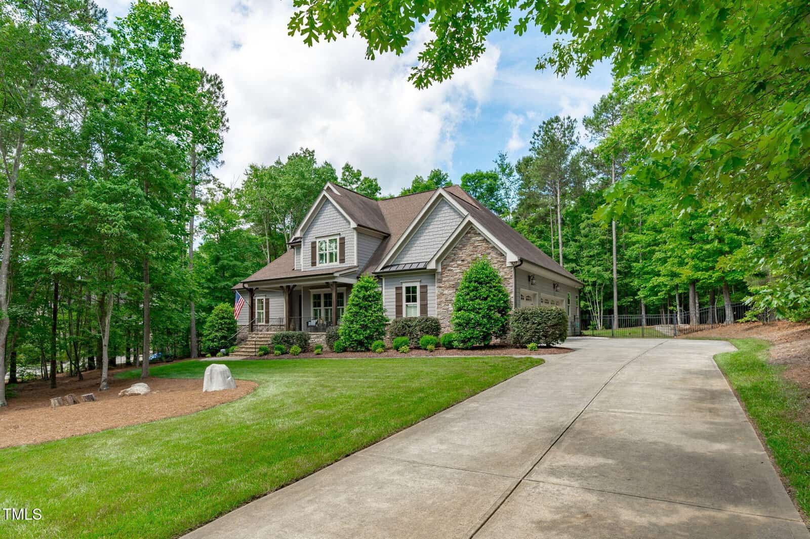 A two-story house with a stone and siding exterior, front porch, and gabled roof sits beside a long curved driveway, surrounded by green lawns, trees, and landscaped shrubs under a partly cloudy sky.