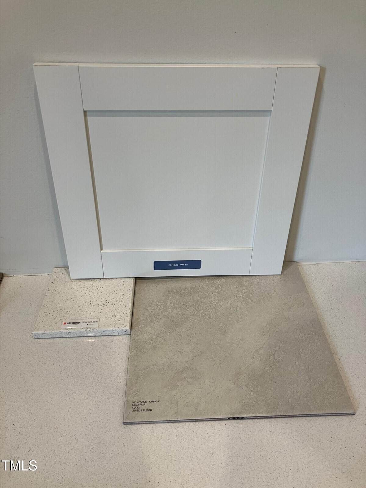 A white shaker-style cabinet door stands upright, with a light gray stone tile and a small, speckled white countertop sample arranged in front of it on a white surface.