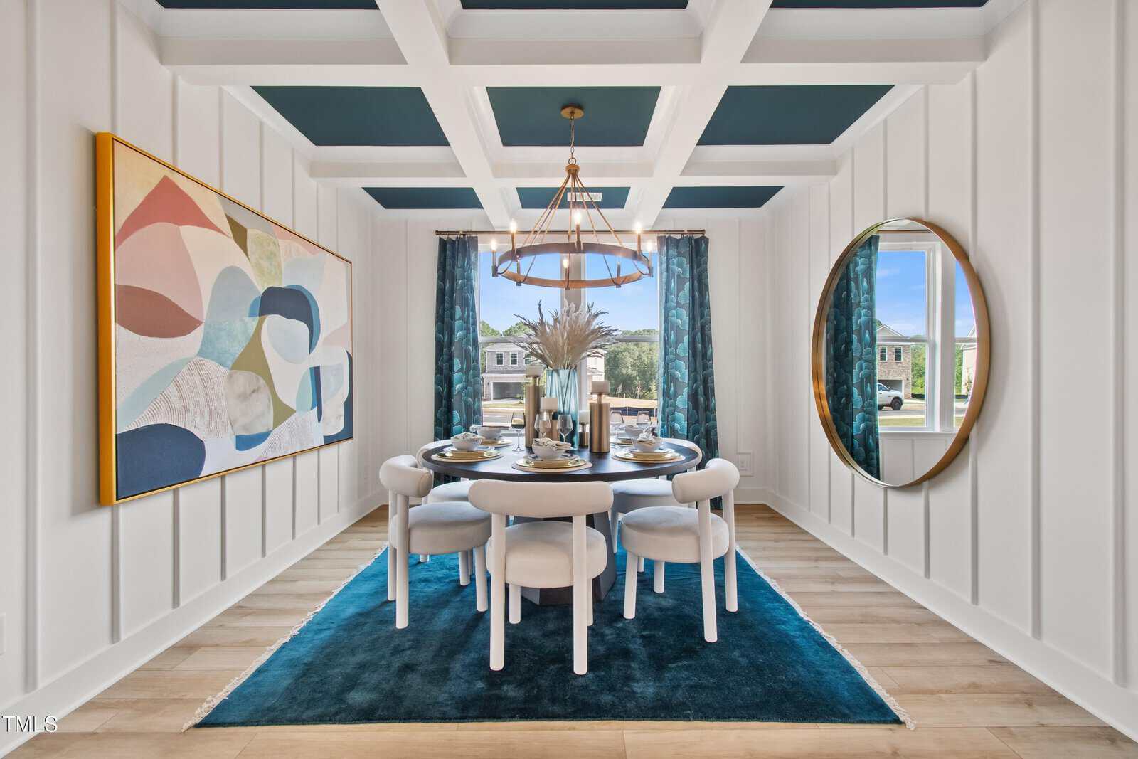 A modern dining room with a round table set for six, white chairs, a blue rug, coffered ceiling, abstract wall art, a round mirror, and large windows with blue curtains letting in natural light.