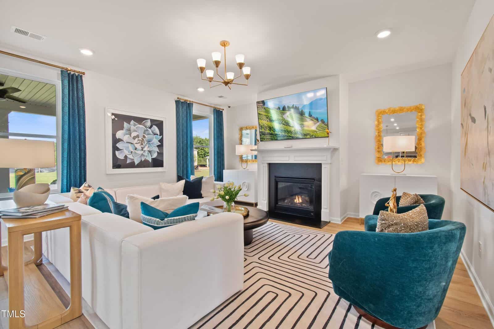 A modern living room with teal and white furniture, a striped rug, a fireplace under a landscape TV, large floral art, gold accents, and blue curtains. Windows show a green outdoor area. Room is brightly lit.