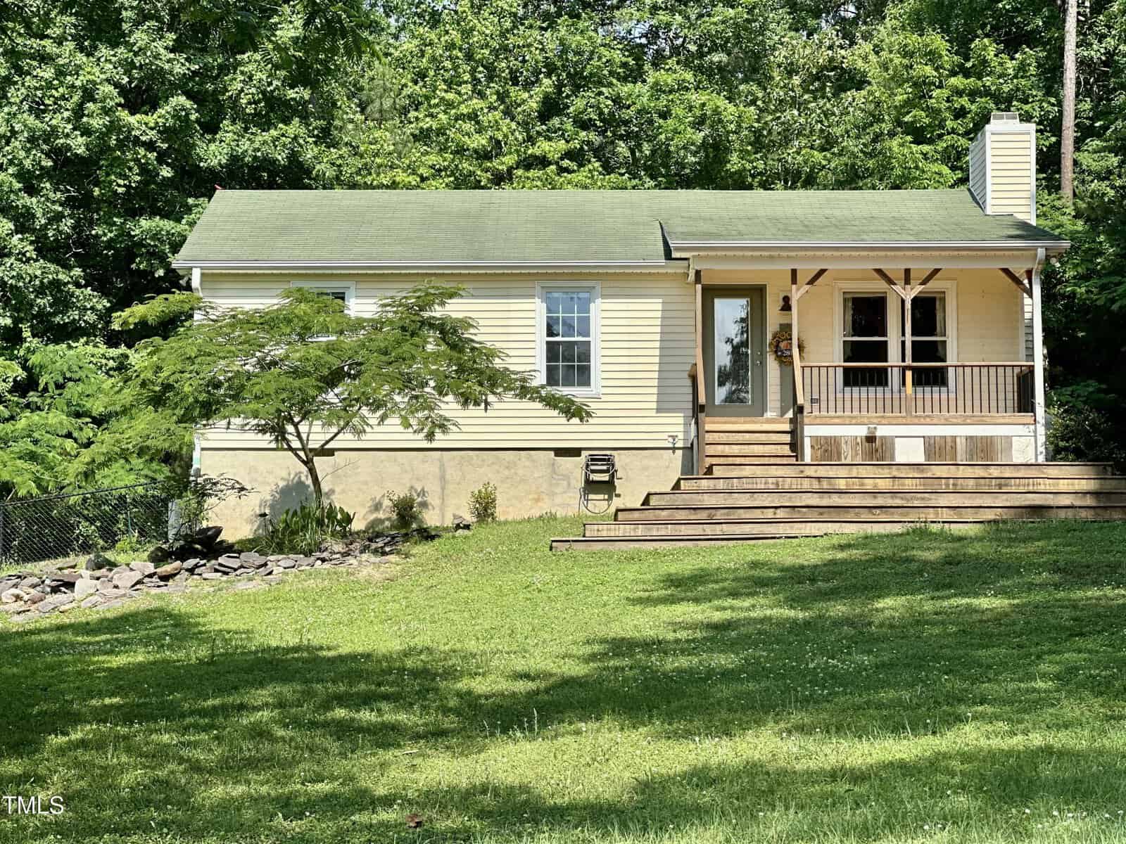 A small yellow house with a green roof and a covered front porch sits on a grassy lawn, surrounded by greenery and trees, with steps leading up to the front door.