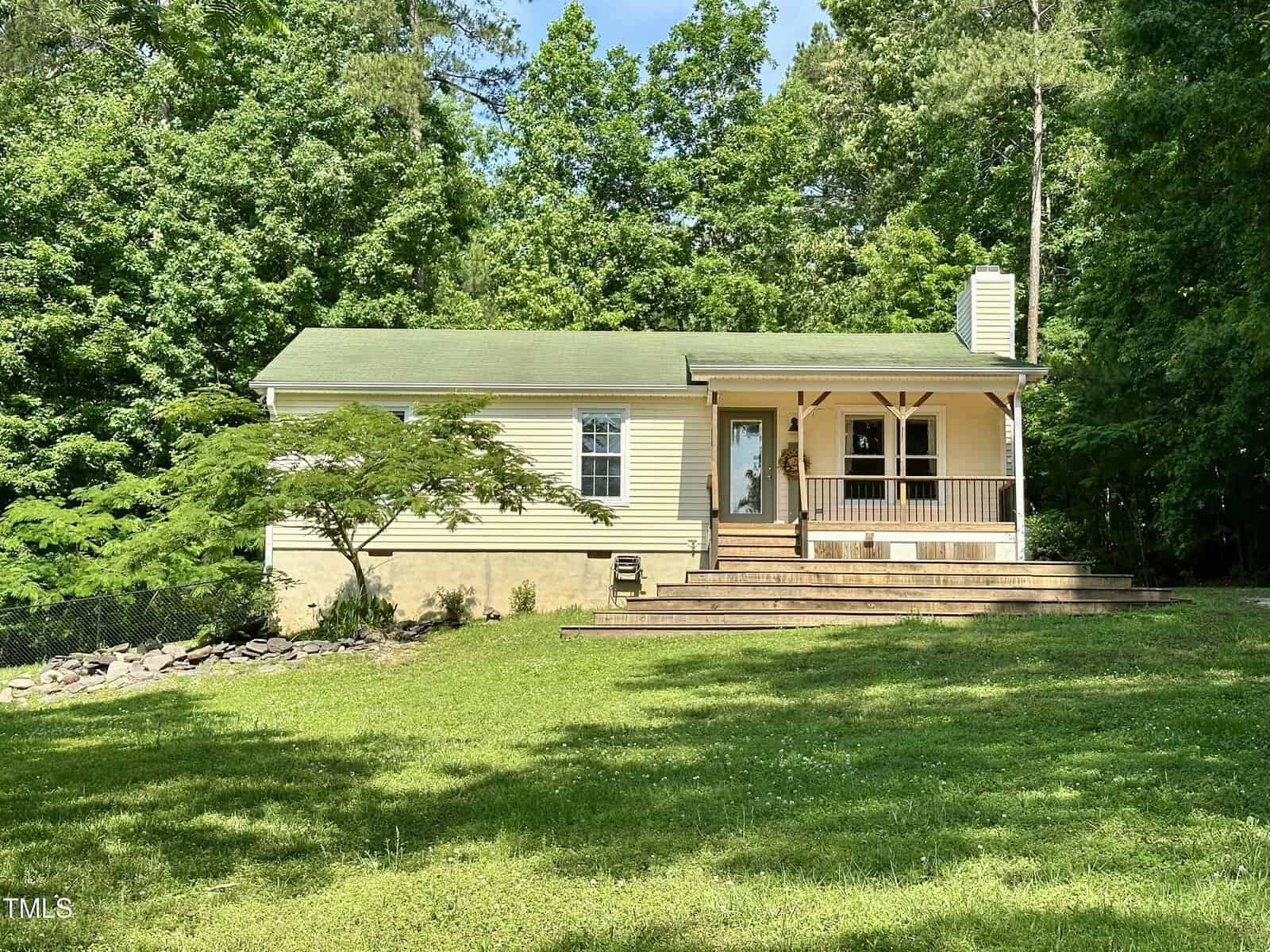 A small, light yellow house with a green roof sits among tall trees. It has a covered front porch with wooden railings and a wide front lawn with some shade from surrounding foliage.