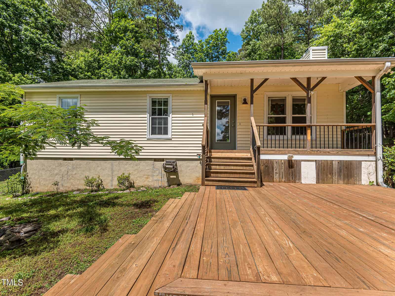 Single-story beige house with a wooden front porch and railing, surrounded by trees and greenery. A wooden ramp leads to the front door. The home has white trim and a stone foundation.