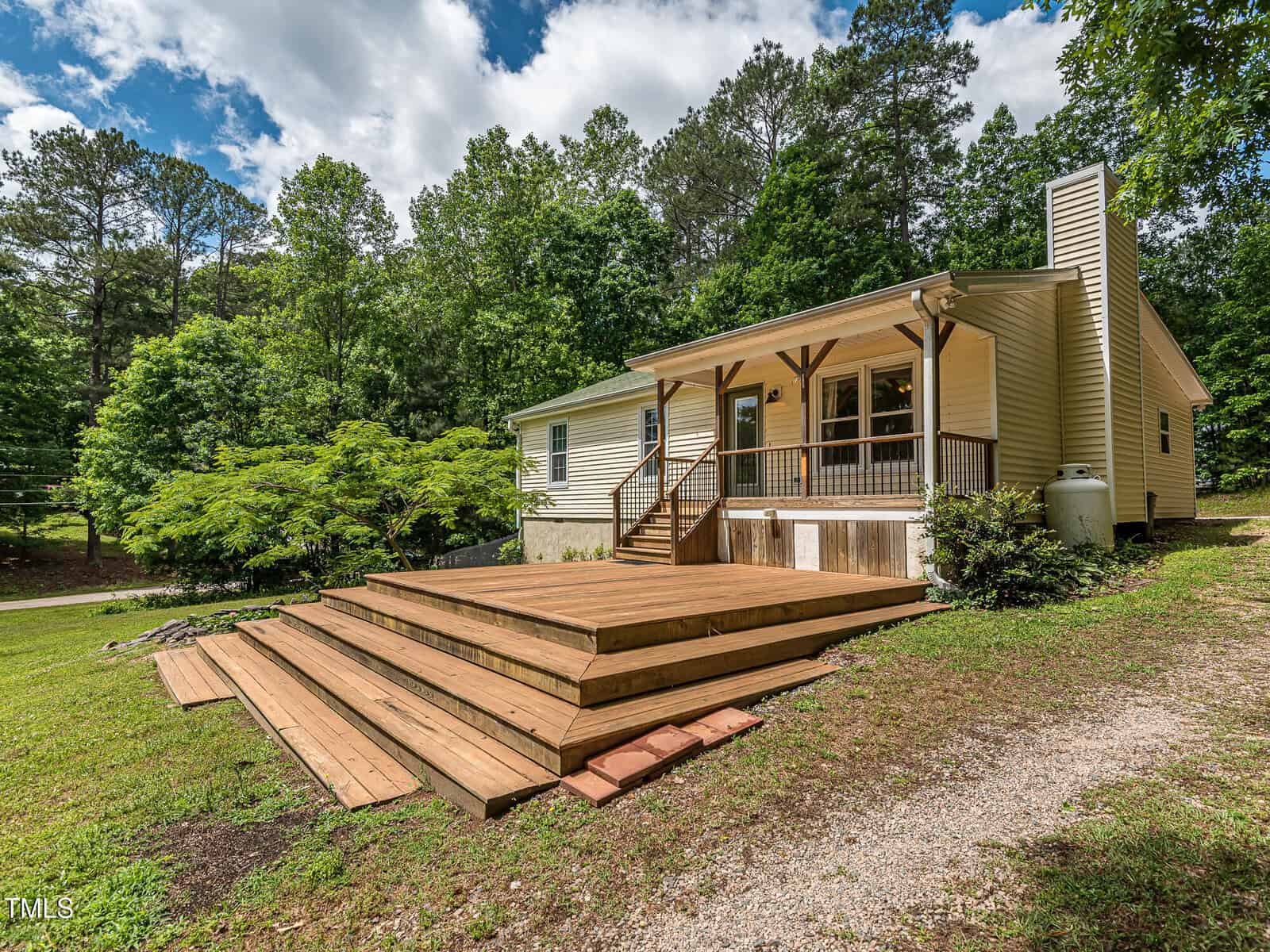 A single-story house with light siding and a large wooden deck surrounded by trees and greenery under a partly cloudy sky. Gravel path and manicured lawn lead up to the home.