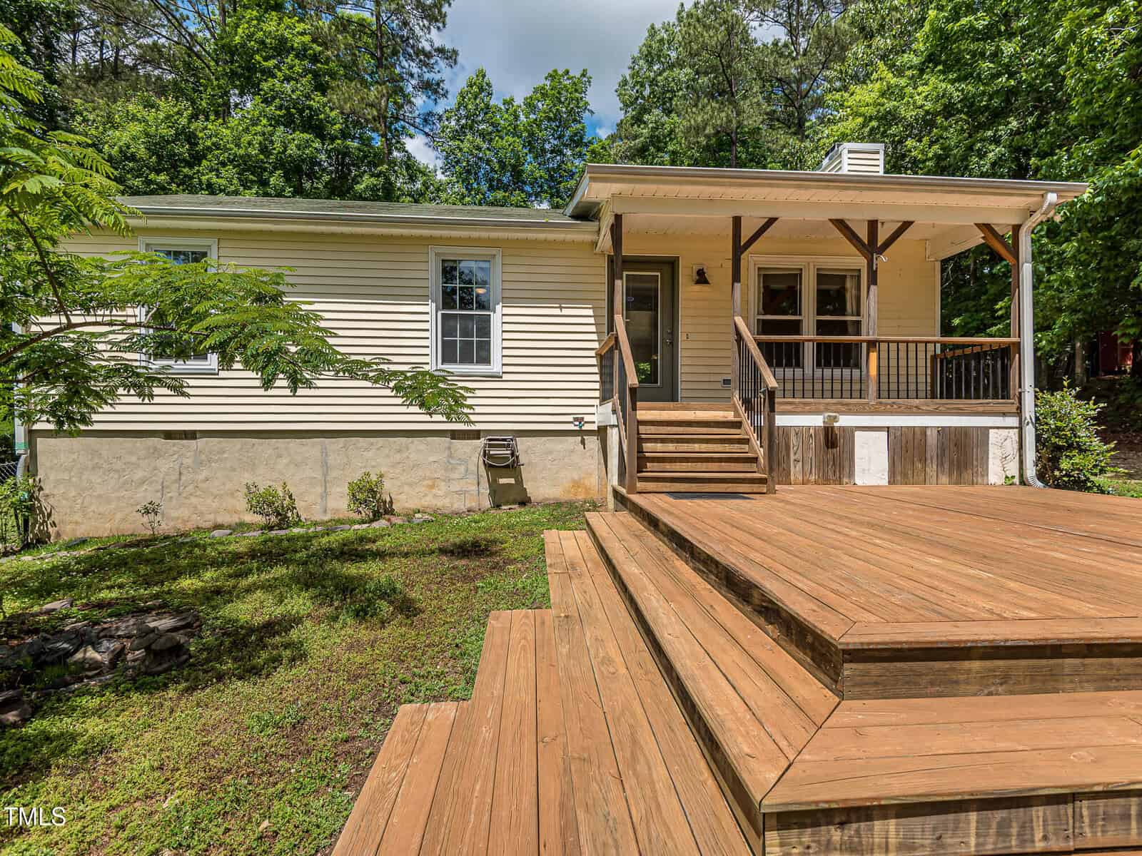 A single-story house with light-colored siding, a covered front porch, and wide wooden steps leading up from a grassy yard, surrounded by trees and greenery.