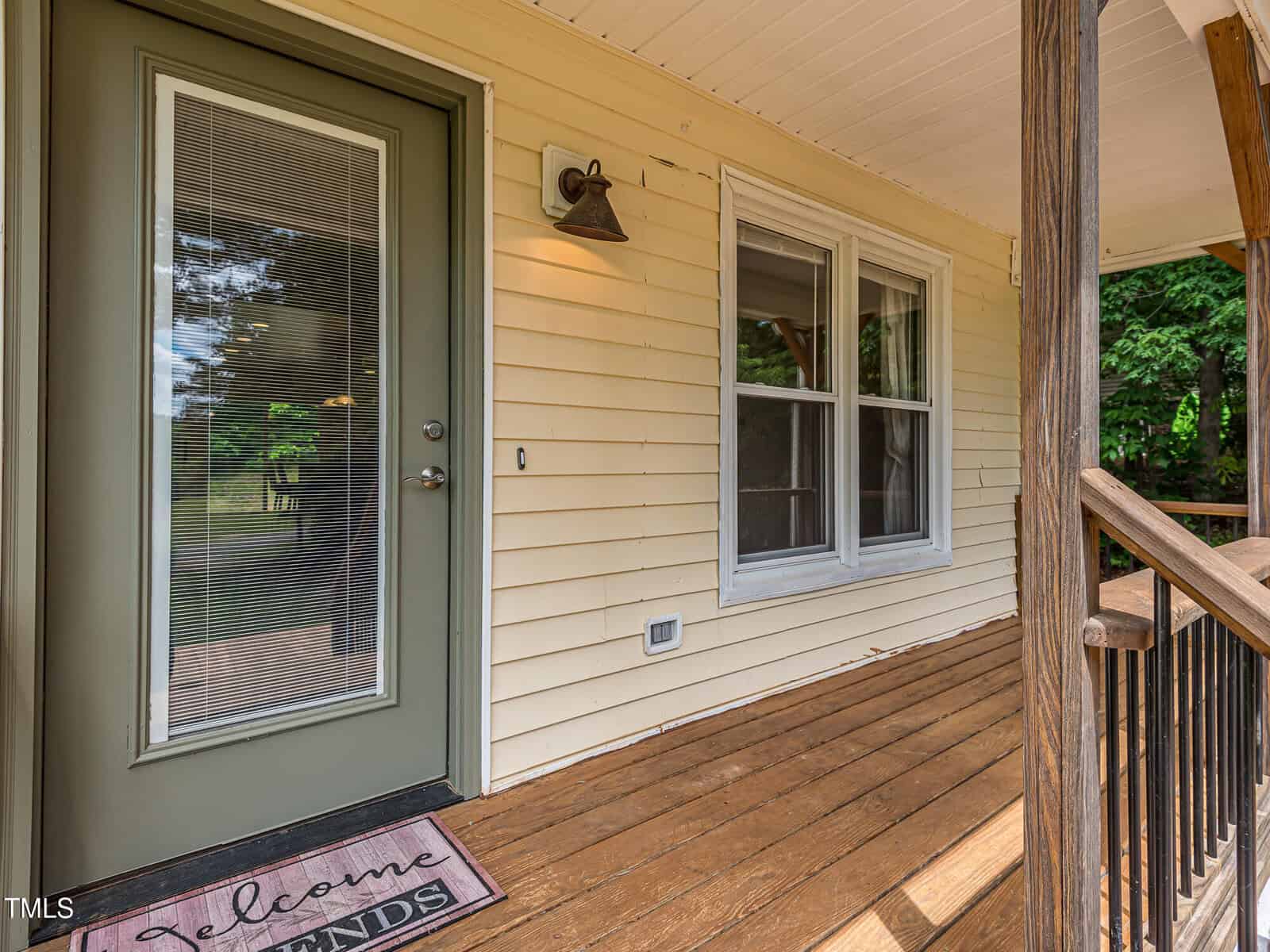 A front porch with wooden flooring and railings, a green door with blinds, a welcome mat, beige siding, an outdoor wall light, and two windows overlooking a grassy yard with trees.