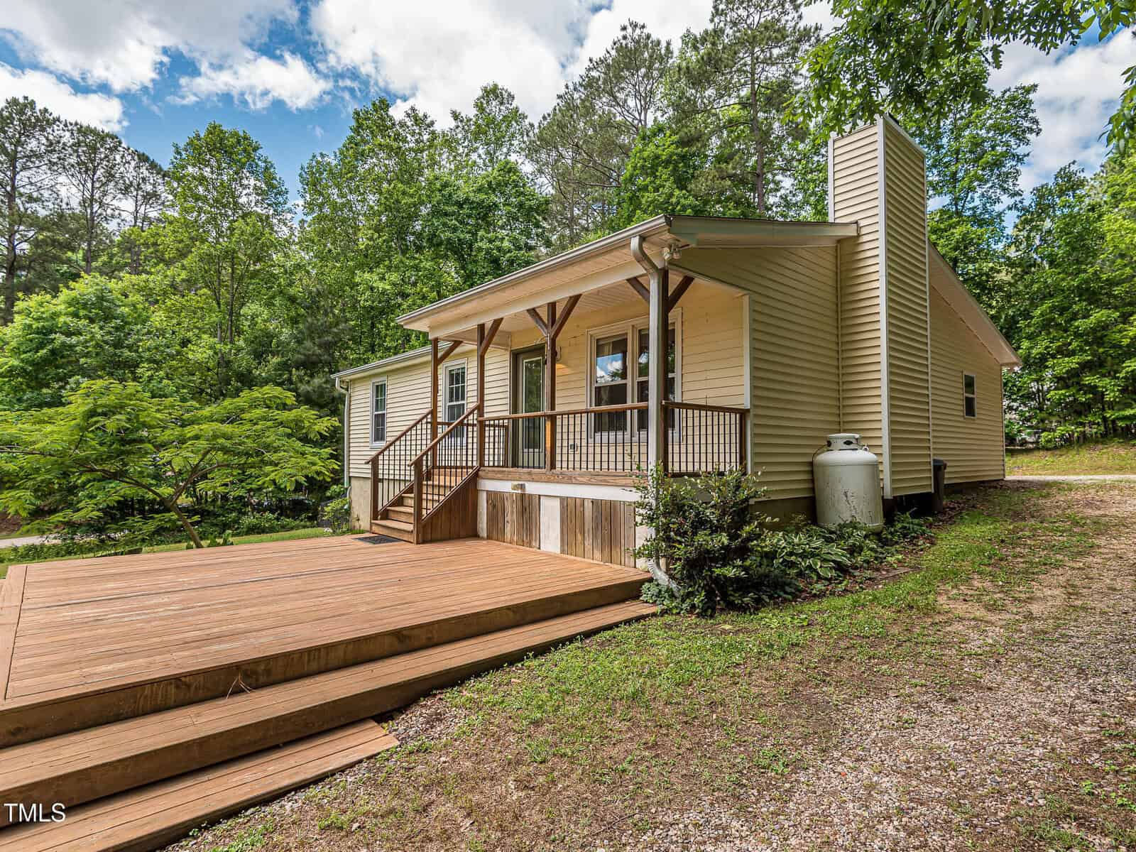 Single-story beige house with a covered front porch, wooden railings, and a large wooden deck. Surrounded by trees and greenery, with a gravel driveway on the right side. Sky is partly cloudy.