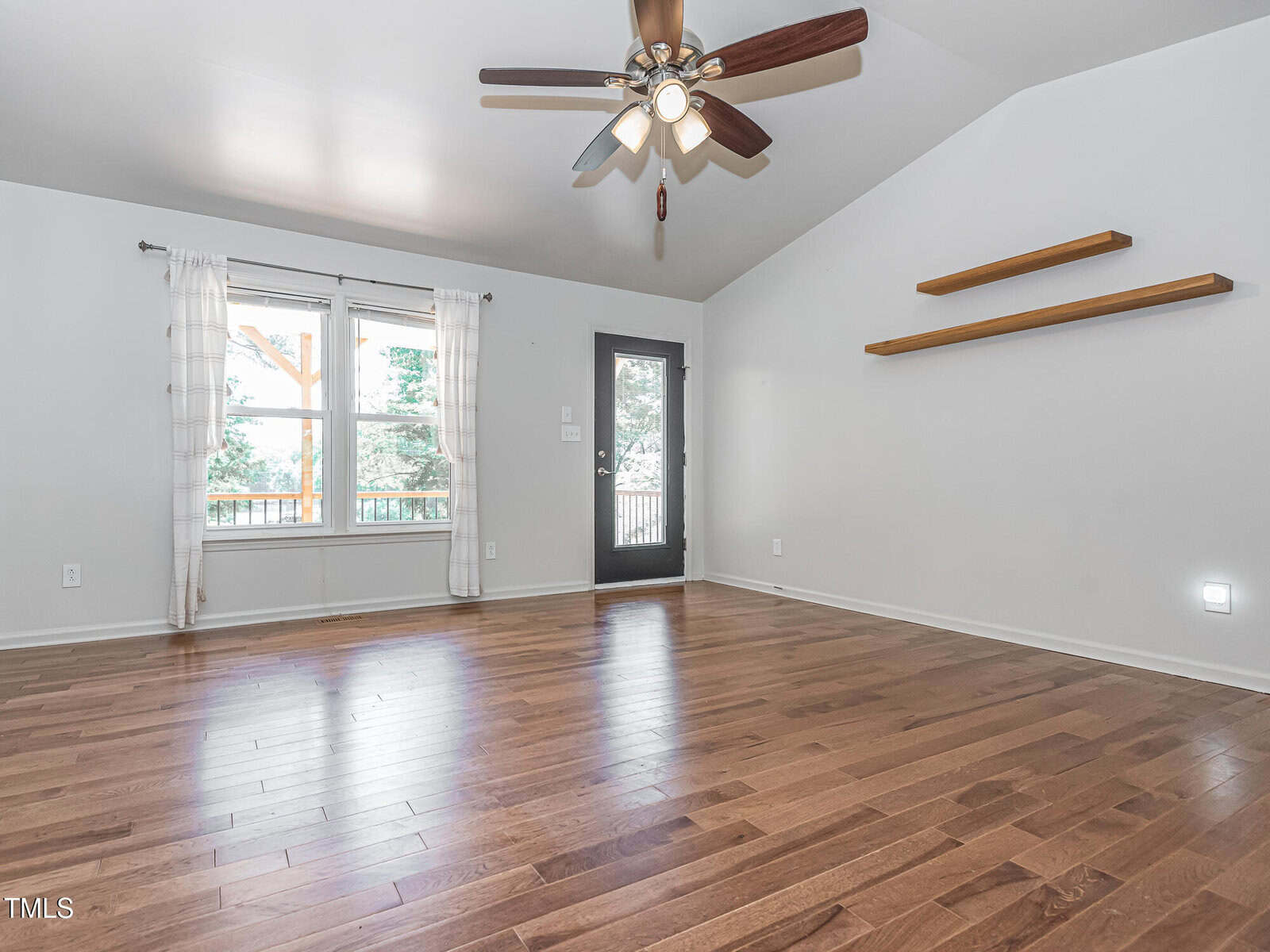 Empty room with light-colored walls, wood flooring, a ceiling fan with lights, two wall-mounted shelves, a window with sheer curtains, and a black door leading outside. Natural light enters through the window and door.