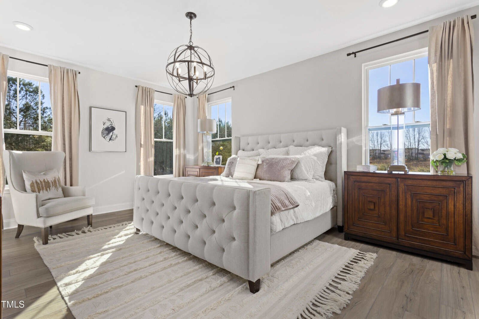 Bright bedroom with a tufted gray bed, striped rug, two wooden nightstands, matching lamps, and neutral decor. Three windows with beige curtains allow natural light in. An armchair sits in the corner near framed wall art.