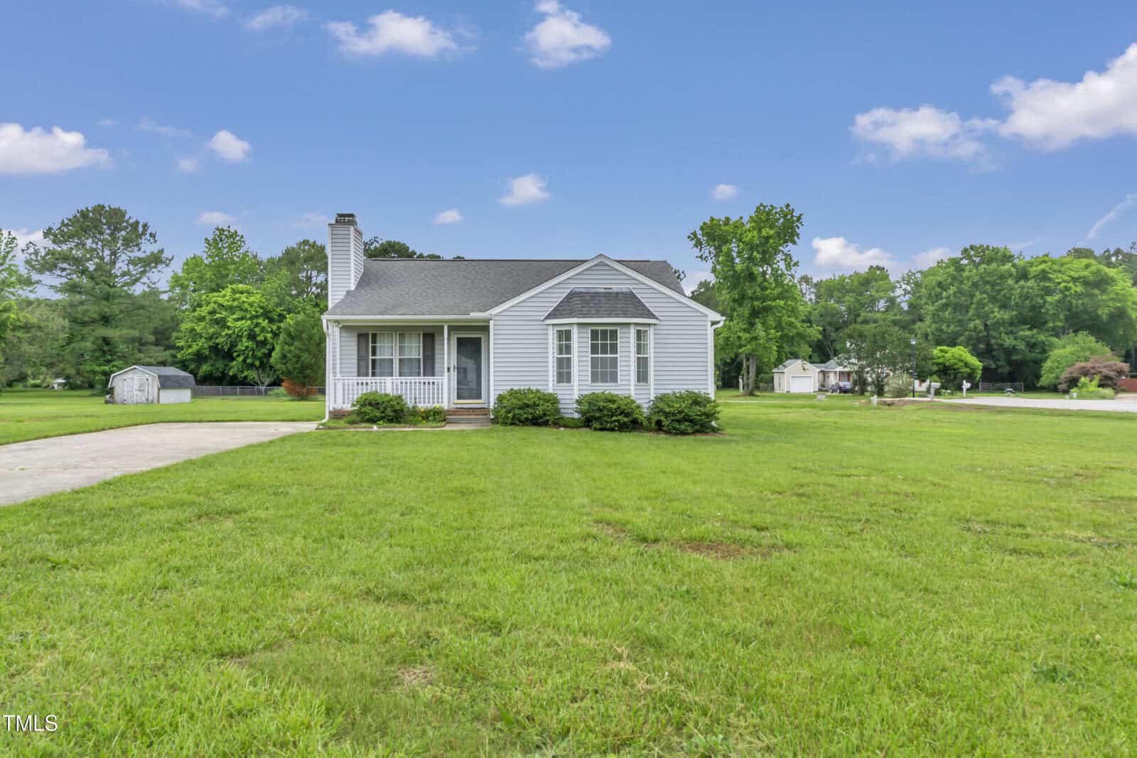 A small, light gray single-story house with white trim sits on a large, neatly mowed lawn under a blue sky with scattered clouds. Trees and a shed are visible in the background.