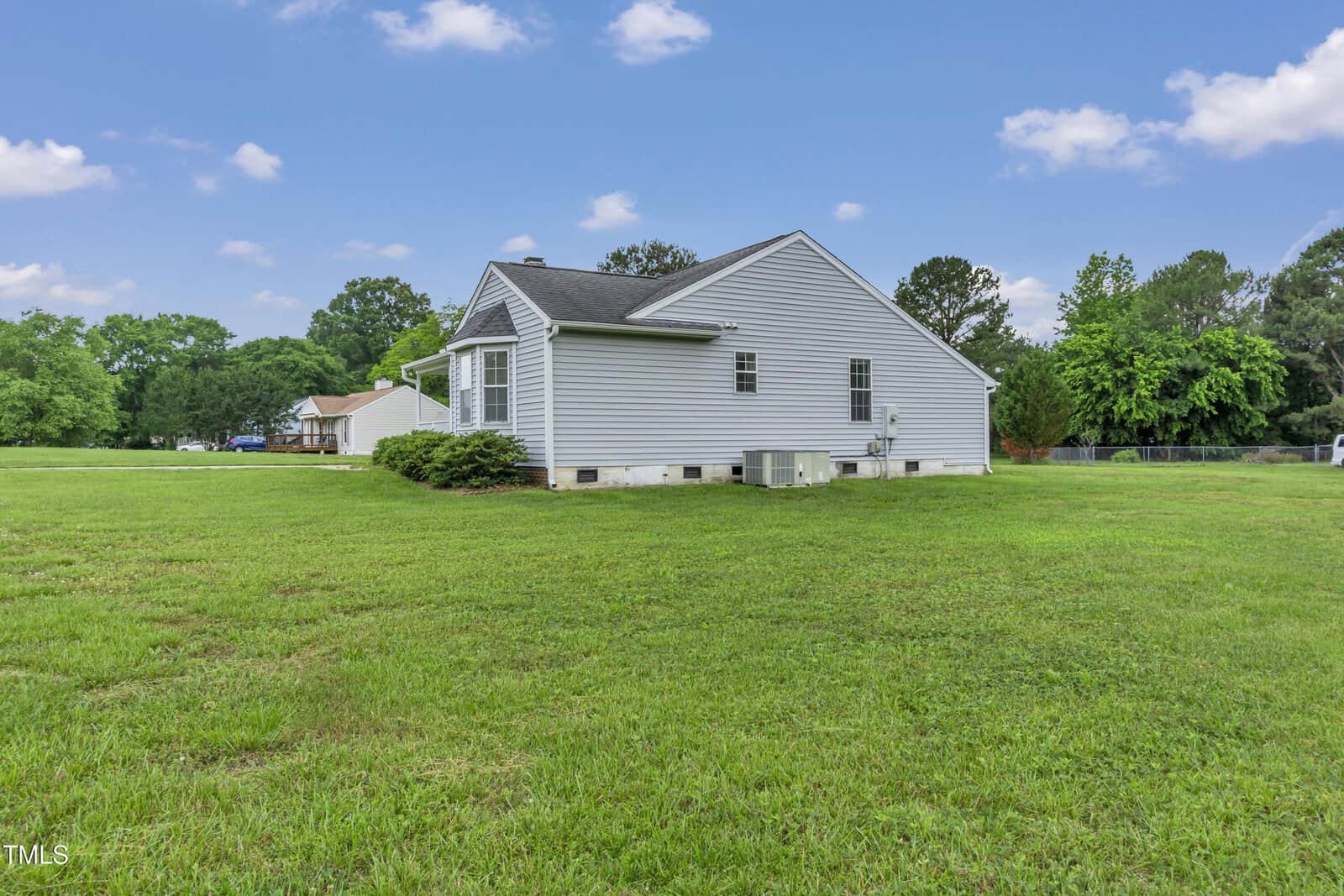 Single-story gray house with white trim, set on a large, grassy lawn. The house has a black roof, several windows, and a visible HVAC unit on the side. Trees and neighboring homes are in the background under a blue sky.