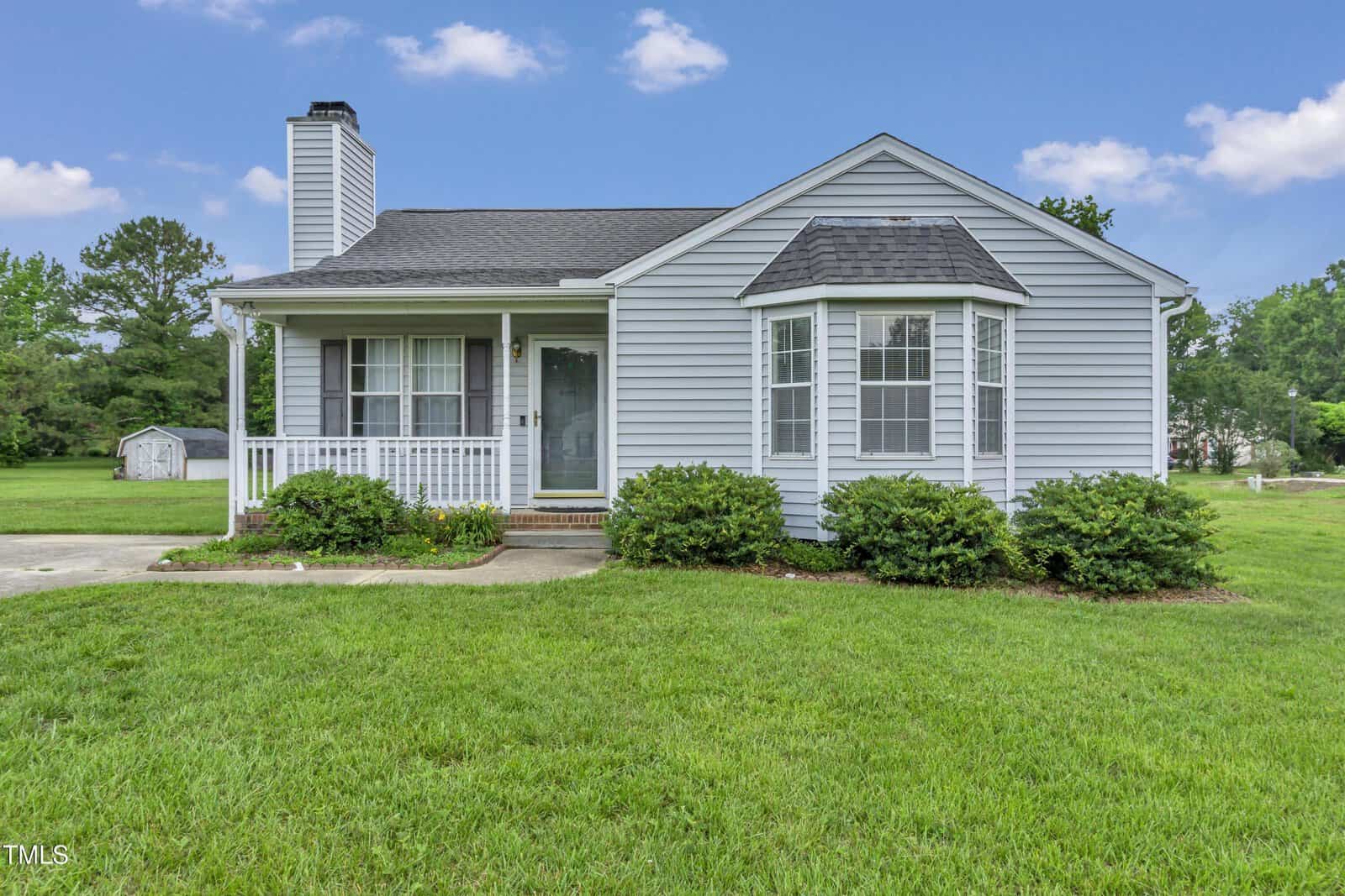 A small, single-story gray house with white trim, a covered front porch, bay window, and neatly trimmed bushes, set on a green lawn with trees and a shed in the background under a blue sky.
