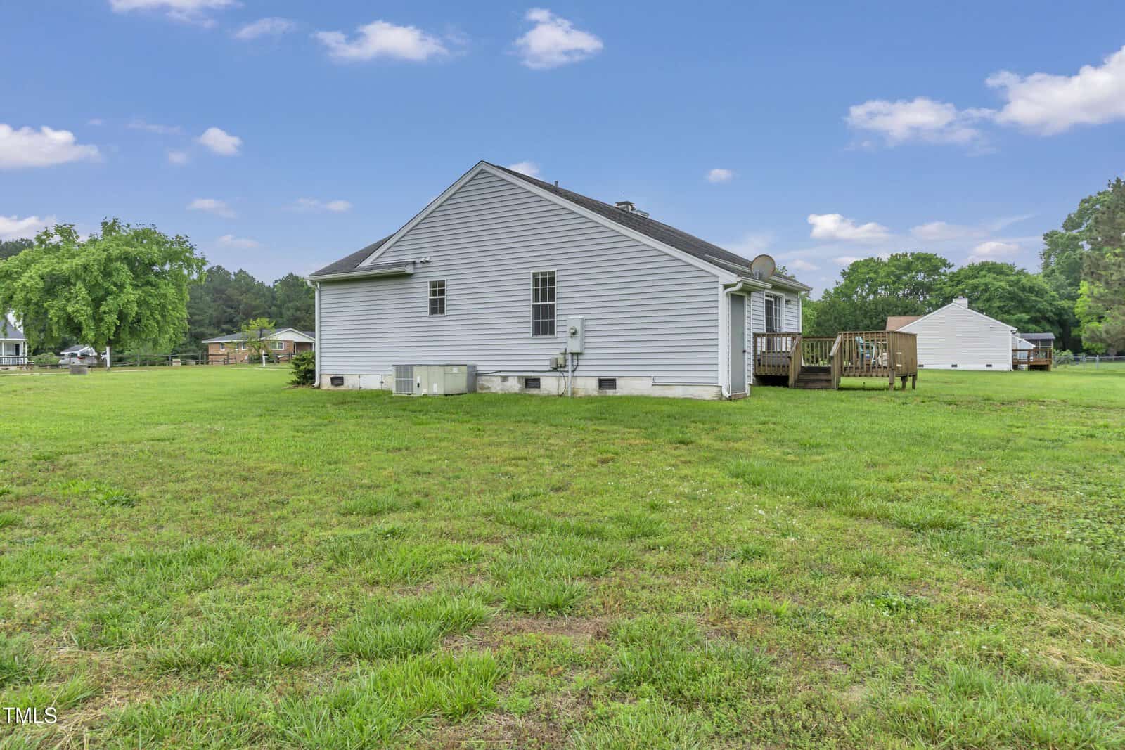 Single-story white house with gray roof, small back deck, and multiple HVAC units, situated on a large grassy yard with scattered trees and a detached white shed in the background under a partly cloudy sky.